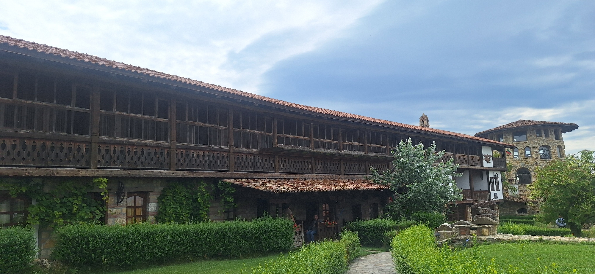 The image displays a large, multi-story building with a long wooden veranda running along its upper levels. The building has a tiled roof and stone lower walls. To the right, a circular stone tower with arched windows rises prominently. Lush green foliage, including trees and hedges, surrounds the structures, with a stone pathway leading through the greenery. The sky is partly cloudy. A sign that reads "SPA" is visible near the pathway. Two individuals are present: one standing by a table on the veranda and another sitting on the edge of the veranda. A third person is standing near the stone tower. The scene is set in the city of Kalimantsi, Bulgaria.
