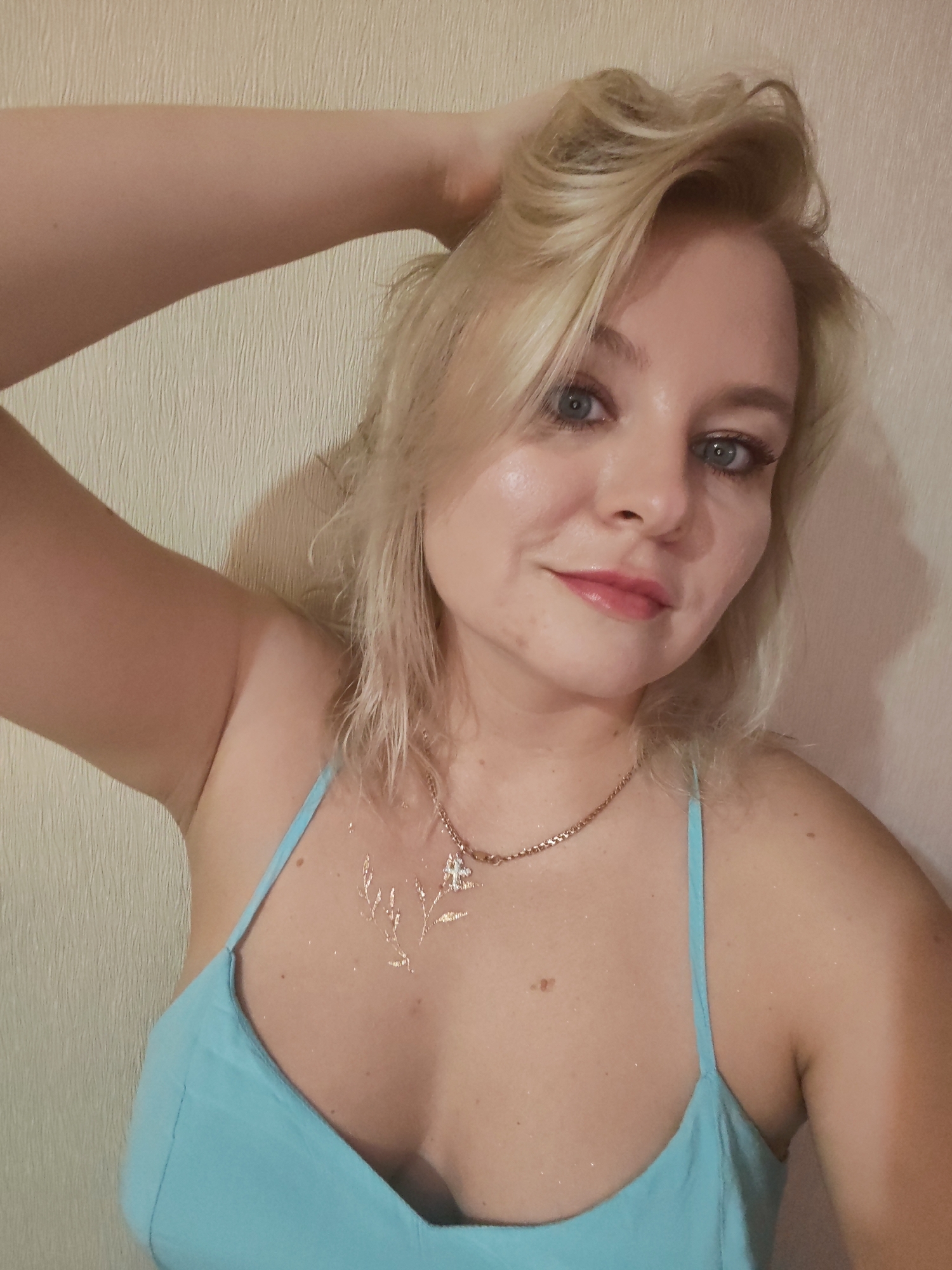 The image shows a young woman with blonde hair and blue eyes, wearing a light blue camisole and a gold necklace. She is posing with her hand in her hair, looking at the camera. There is a faint shimmer on her chest and shoulders, possibly from glitter or body lotion. The background is a textured, light-colored wall. The image does not contain information that allows for identification of a specific city, such as Izhevsk, Russia, or any elements that would be relevant for a forensic analysis.