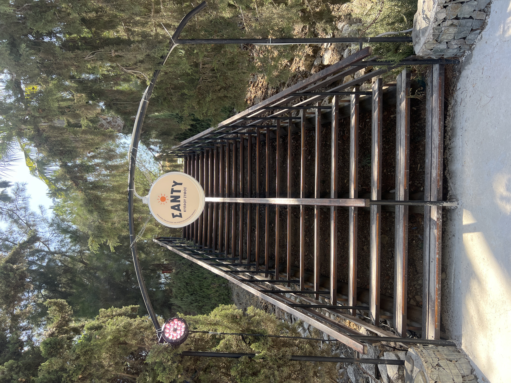 An exterior shot captures a set of wooden stairs with metal railings leading upwards, flanked by trees and greenery. A circular sign hangs above the stairs, featuring a sun graphic and Greek text. To the left of the stairs, a large, circular light fixture with pink LEDs is mounted on a pole. The scene appears to be located outdoors, possibly in a park or at an establishment. The text on the sign reads "ΣΑΝΤΥ" and below that, "ΚΥΠΡΟΥ ΣΥΘΟΣ". This sign likely indicates the name of a place or event. The overall impression is that of an entrance or pathway to a venue, set against a natural, sun-dappled background.