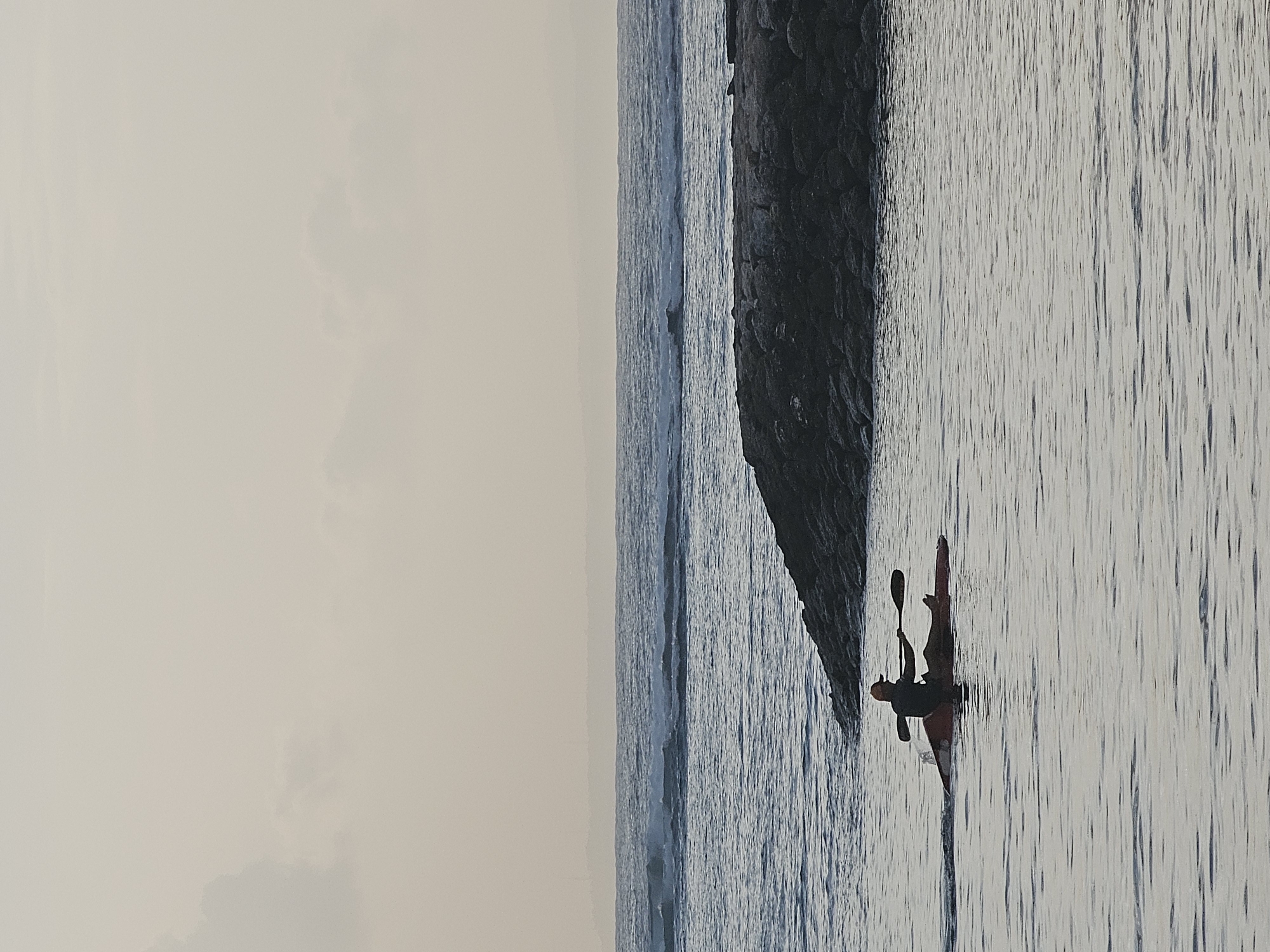 A lone kayaker, silhouetted against the water, paddles a red kayak in the ocean. The kayaker is wearing a hat and appears to be mid-stroke. In the background, waves crash against a rocky breakwater. The sky is overcast with soft clouds. The scene suggests a peaceful, possibly early morning or late afternoon, setting in Denpasar, Indonesia, likely near a beach or coastal area.