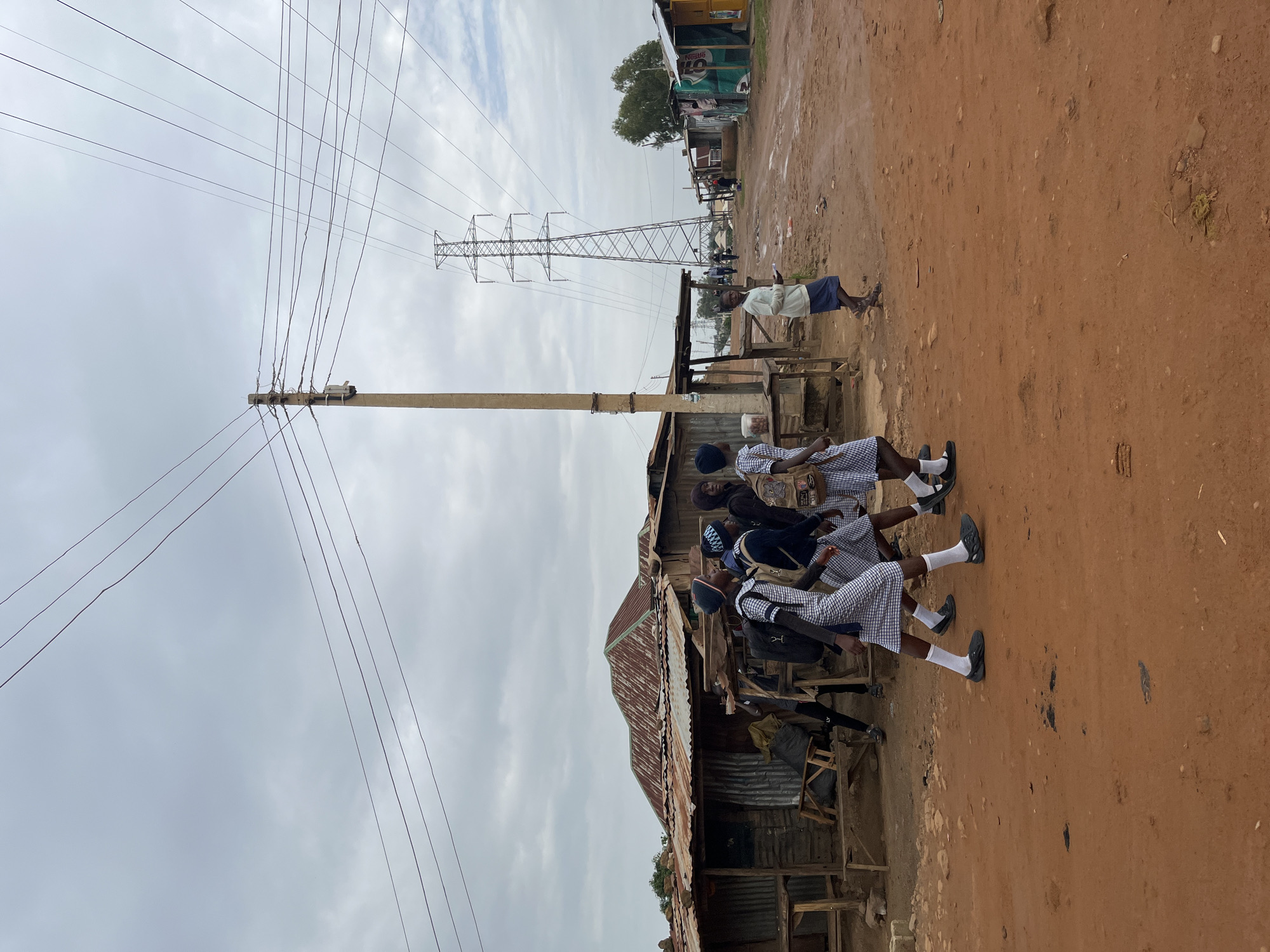 The image depicts an outdoor scene in Jos, Nigeria, with a focus on a dirt road and several structures. In the foreground, four children in school uniforms, two boys and two girls, are walking from left to right. They are all wearing checkered skirts or shorts, white socks, and dark shoes, and some are carrying backpacks. A fifth child, wearing a blue skirt and a white shirt, is walking towards the right from the middle ground.
The environment is characterized by several makeshift structures with corrugated iron roofs, suggesting a densely populated or informal settlement. These structures appear to be shops or residences. A large electricity pylon and several power lines are visible in the background, indicating the presence of electricity infrastructure. The ground is covered in reddish-brown dirt, typical of the soil in the region. The sky is overcast and appears cloudy. The overall scene suggests a daily life activity in a developing urban or semi-urban area of Jos, Nigeria.