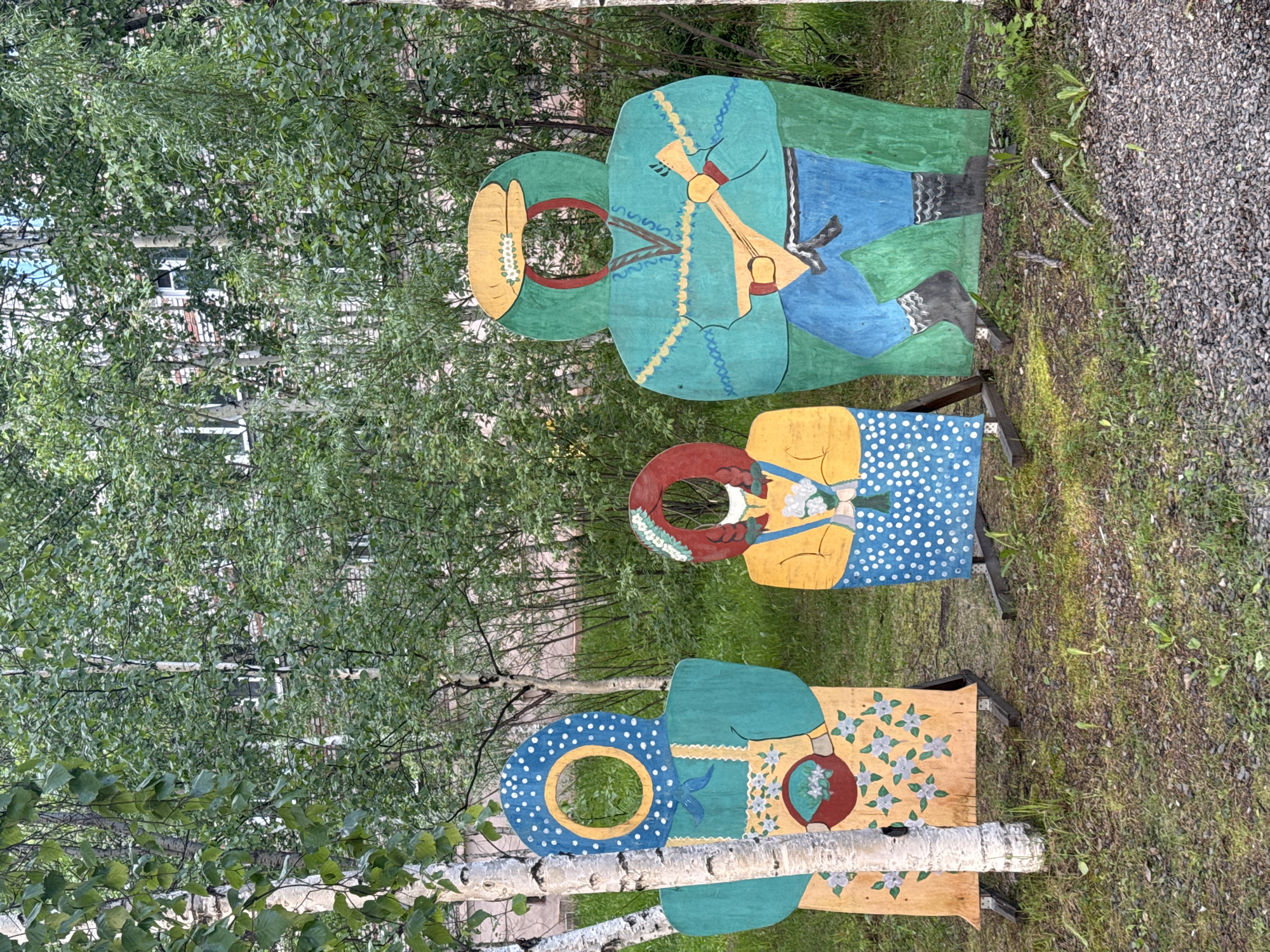 The image shows three wooden cutout figures with holes for faces, placed outdoors in a grassy area with trees. The figures are painted to represent people in traditional clothing. The leftmost figure is a woman wearing a blue headscarf with white polka dots, a green apron over a yellow dress with floral patterns, and is holding a basket of flowers. The middle figure is a smaller woman wearing a red head covering, a yellow top, and a blue polka dot dress, holding a bouquet of flowers. The rightmost figure is a man in a green hooded garment over blue pants and black boots, playing a stringed instrument. The backdrop consists of lush green trees and foliage, with a hint of a brick building visible through the leaves. The overall setting suggests a park or an outdoor recreational area in the city of Tynda, Russia, likely intended for tourists or visitors to pose for photographs.
