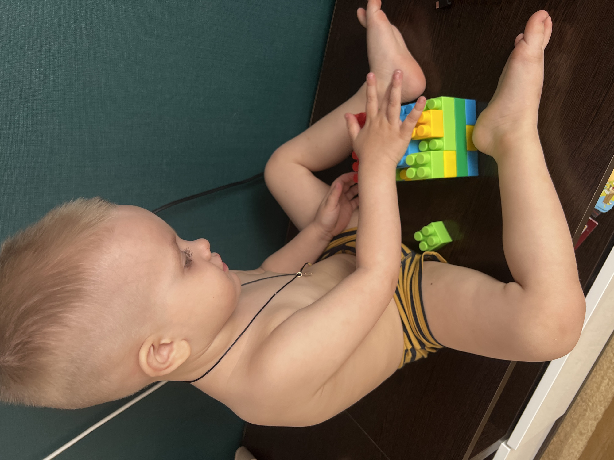 A toddler with blond hair is lying on a dark brown surface, possibly a table or desk. The child is wearing striped yellow and black underwear and a thin black necklace. The child is playing with colorful plastic building blocks in shades of green, yellow, and blue. The toddler is lying with feet extended and hands interacting with the blocks. A green textured wall is visible in the background. The location is Марковский, Russia.
