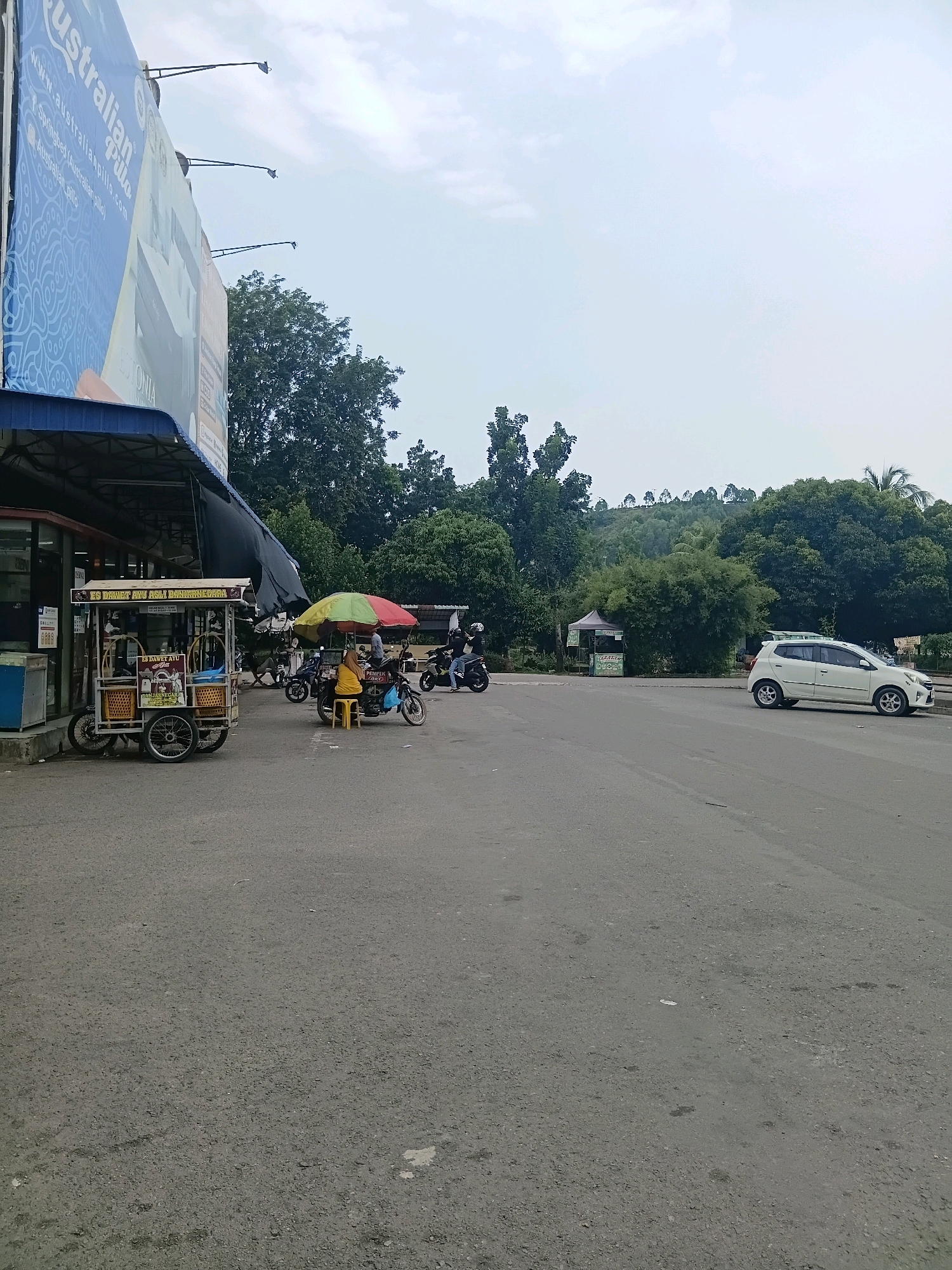 The image depicts a street scene in Batam, Indonesia, likely near a commercial area with a billboard, food stalls, and a road. On the left, a large billboard advertising "Australian Pillow" is visible, supported by metal frames. Adjacent to the billboard is a food stall with a blue awning, selling what appears to be iced drinks. In front of the stall, a wheeled cart with baskets and a sign reading "ES DAWET AYU" is present. Further to the right, a person is sitting on a yellow stool under a multi-colored umbrella, with a food cart nearby selling "PEMPEK PALEMBANG". Several motorcycles are parked around this area, and two people are on one of the motorcycles, with one person wearing a black helmet and the other a white helmet. A white car is parked on the right side of the road, facing away from the camera. The background shows lush green trees and a hilly landscape under a cloudy sky. The road surface is asphalt, and the overall impression is that of a typical day in a bustling area of Batam.