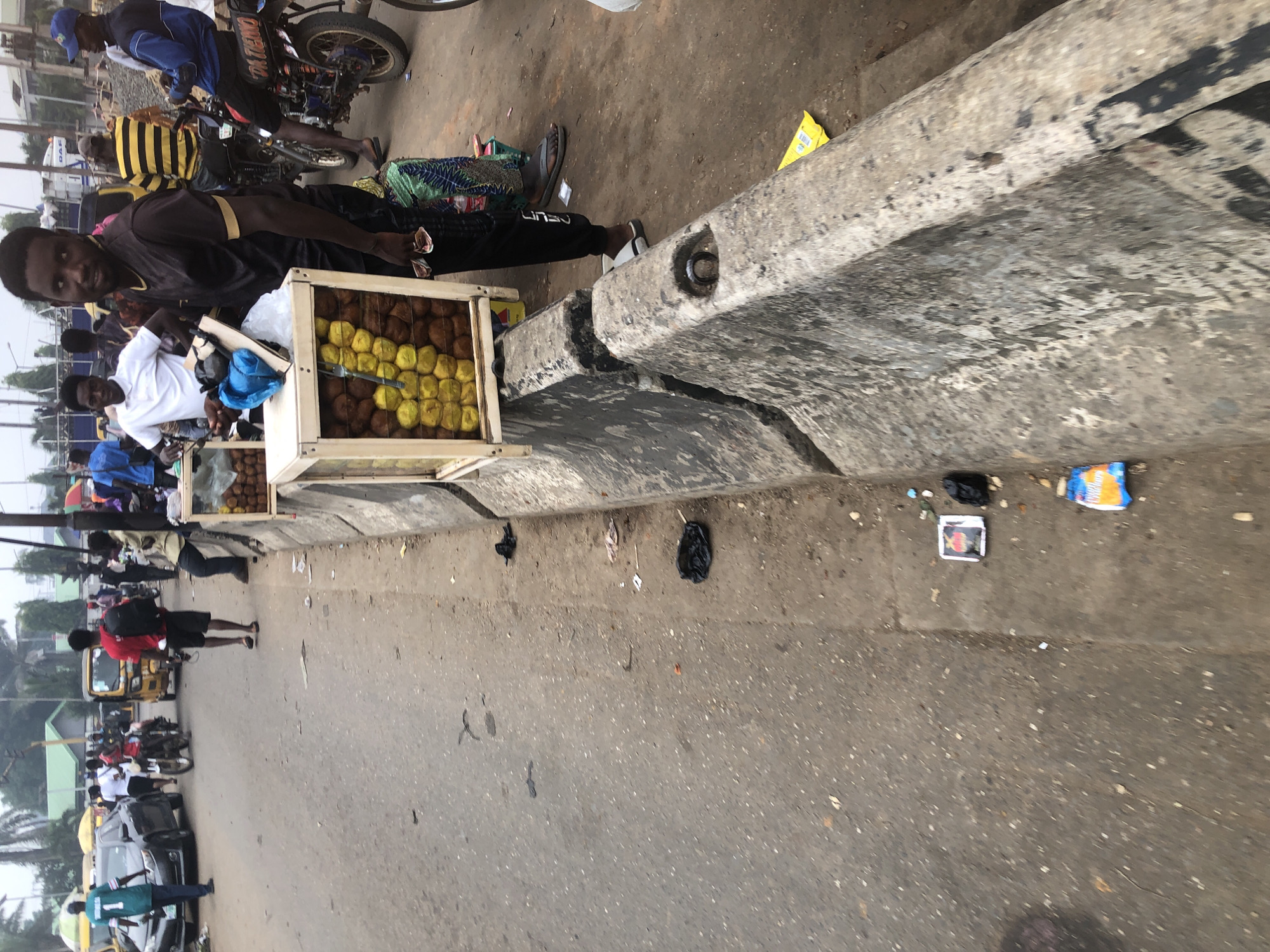 The image captures a bustling street scene in Isheri, Nigeria. A young man, wearing a black and gold shirt and black pants, stands beside a wooden and glass display filled with yellow and brown spherical objects, likely fruits or snacks. He is holding some items in his hand and looking towards the camera. Behind him, other people and motorcycles are visible, suggesting a busy marketplace or roadside vending area. The street is paved with asphalt, and a concrete barrier runs along the side, littered with some debris. In the background, buildings and trees are present, indicating an urban or semi-urban environment. The overall impression is one of everyday life and commerce in a Nigerian city.