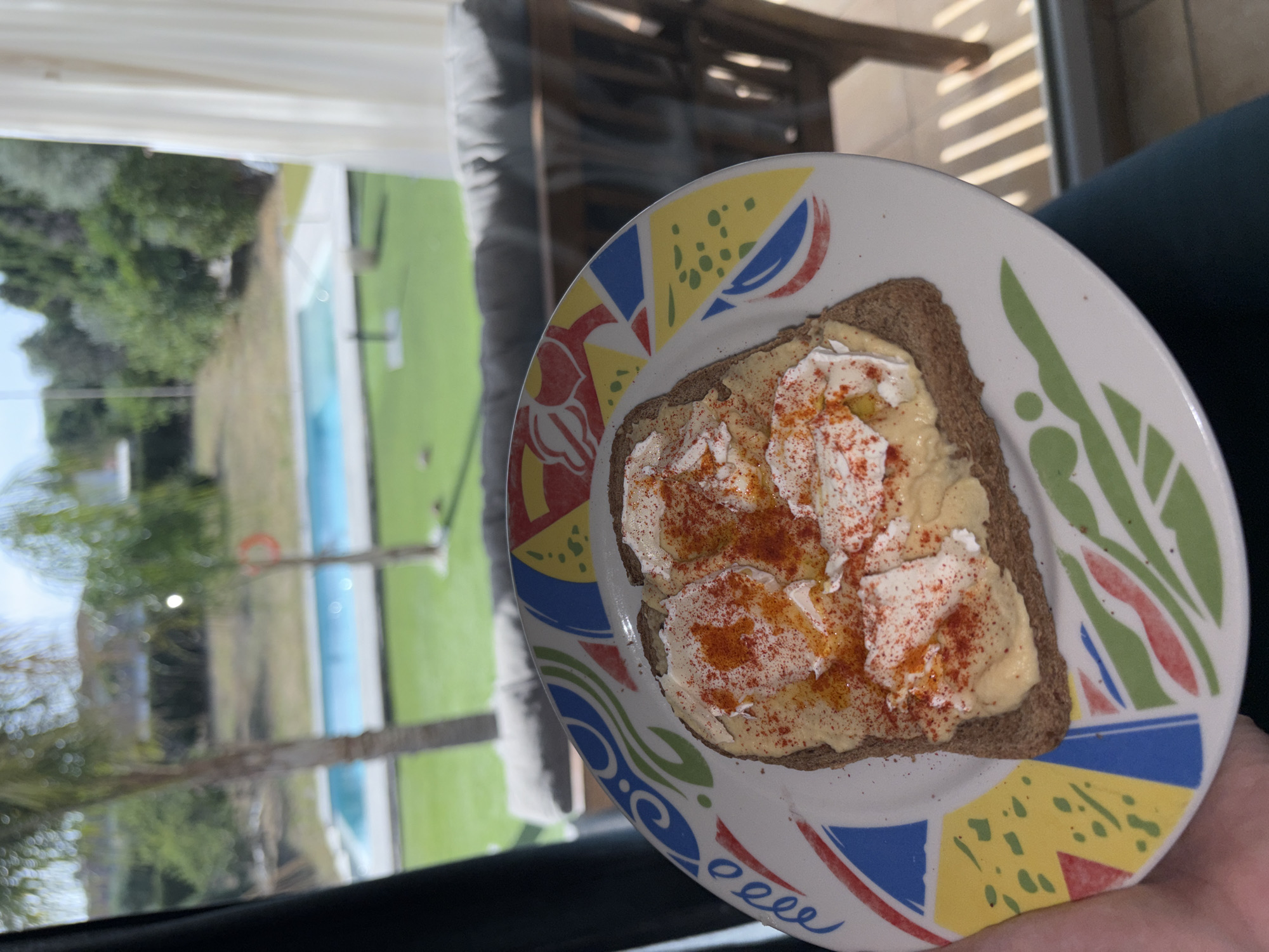 The image shows a piece of whole-wheat toast on a white plate with a colorful pattern. The toast is spread with a yellow substance, possibly hummus or a similar spread, and topped with dollops of white cheese or cream cheese, sprinkled with red paprika. The plate is held by a person. Through a window, a blurred view of an outdoor area is visible, including a swimming pool, green grass, and trees, suggesting a residential setting. The location is Urbanización Encinares De Alcolea, Spain.