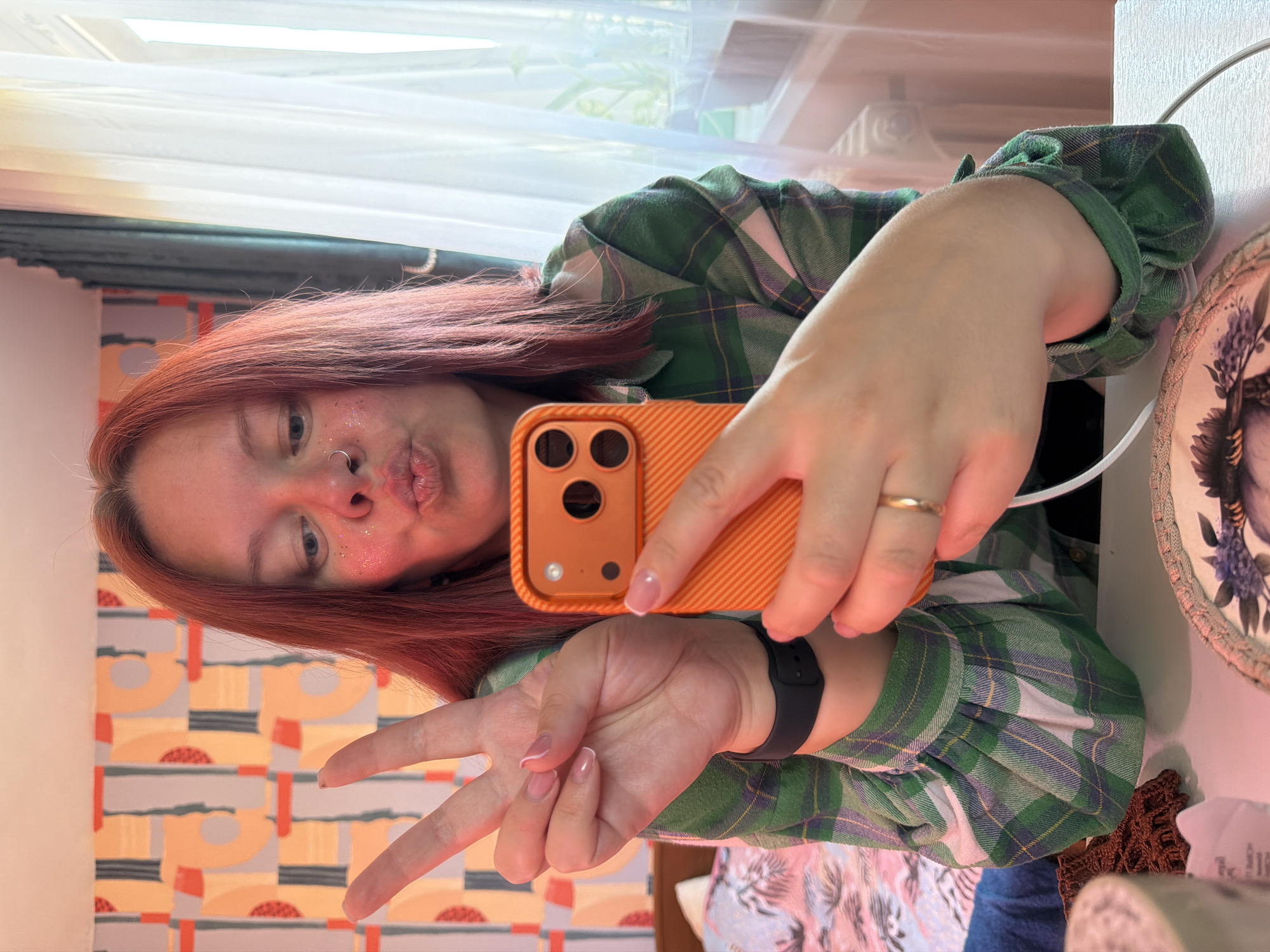A woman with red hair is taking a selfie indoors. She's wearing a green and white plaid shirt and holding an orange phone. She's making a "peace" sign with one hand and puckering her lips for the photo. She is wearing a ring on her finger and a smart watch. The background includes a patterned wall, a window with light curtains, and a round decorative plate. Based on the context of the task, this is in Chaikovsky, Russia.