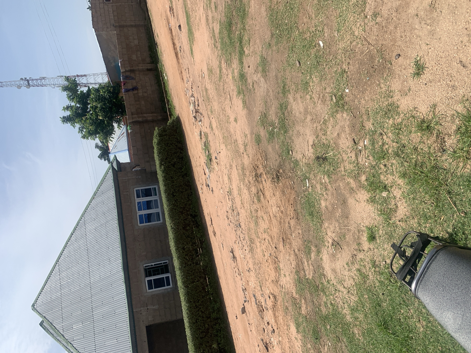 The image shows an outdoor scene in Jauro Yinu, Nigeria. In the foreground, a portion of a dark, textured seat and a black metal rack, likely from a motorcycle or bicycle, are visible on the left side. The ground is a mixture of reddish-brown dirt and sparse green grass. In the middle ground, a well-maintained, dense green hedge runs horizontally across the frame, separating the foreground from a building and a wall. The building behind the hedge has a light brown, brick-like facade and a corrugated metal roof. It features two white-framed windows with blue panes. Beyond the building and hedge, a wall constructed of grey, rectangular blocks stretches across the background. To the right of the building, a tall, white telecommunications tower with multiple antenna structures is visible against a bright, pale blue sky with some wispy white clouds. A green tree with dense foliage stands to the left of the tower. Some clothes are hanging on the wall. The overall environment appears to be a rural or semi-urban setting.
