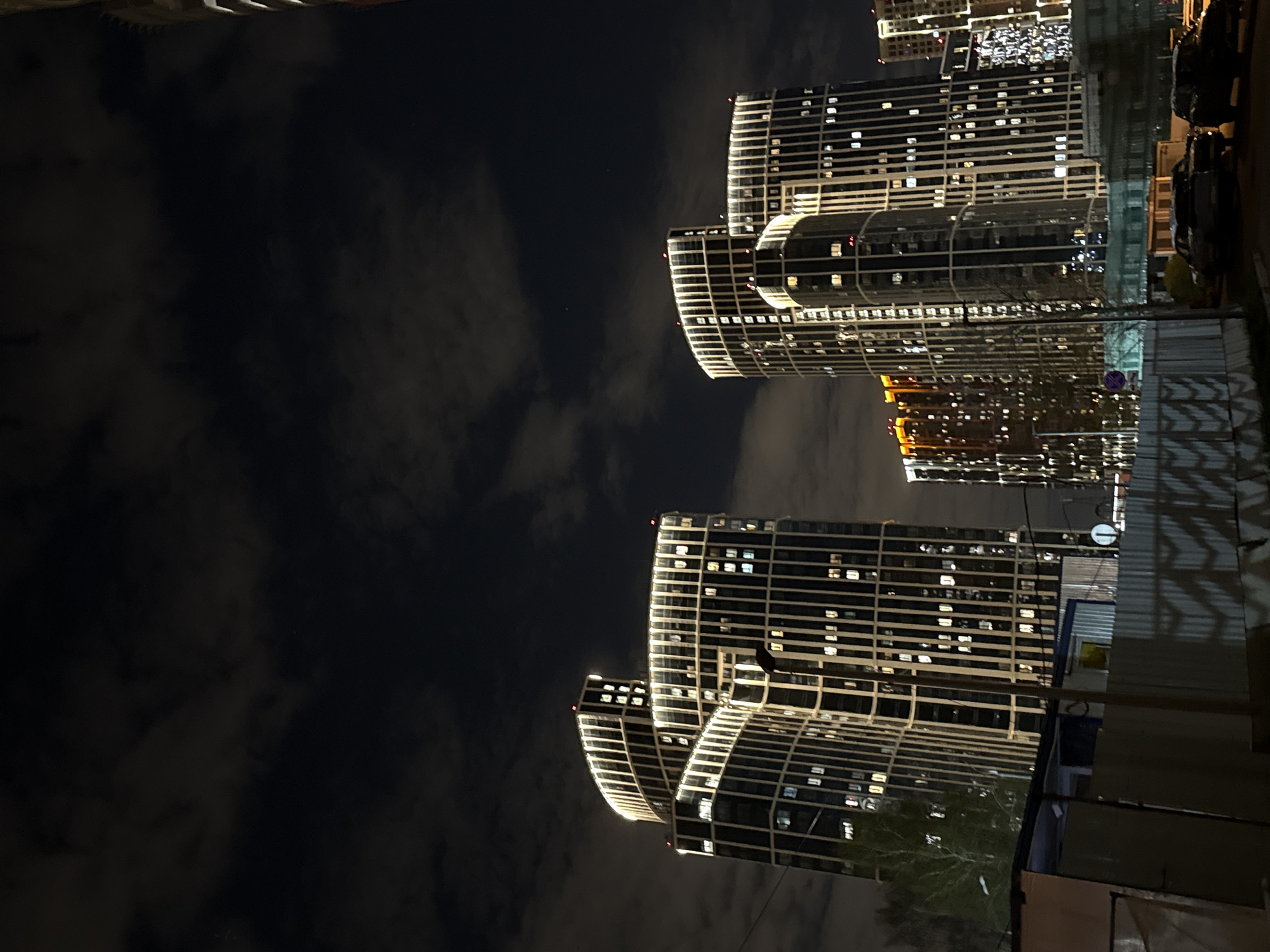 This nocturnal image captures a modern cityscape, appearing rotated approximately 90 degrees clockwise from a standard landscape orientation. The scene is dominated by several contemporary high-rise buildings, illuminated brightly against a dark, cloudy night sky.
The prominent subjects are the architectural structures, characterized by extensive external lighting that outlines their shapes with horizontal and vertical light strips. Numerous windows are visible, many glowing from within, indicating occupancy. Some buildings display sleek, curved designs, contributing to their modern aesthetic. The sky is dark with discernible, softly lit clouds, suggesting a cloudy but clear night.
Towards the bottom and right edges of the rotated image, elements of the street level are visible, including dark outlines of parked vehicles, what appears to be a street lamp, and possibly construction barriers or fences. A faint, blue circular sign, likely a traffic sign, is barely visible in the mid-ground. Bare tree branches are also present near the base of some buildings.
No specific activity or event is taking place; the image presents a static view of an urban environment at night. There is no legible text visible within the frame. The overall impression is one of a vibrant yet quiet urban setting, consistent with a city like Khimki, Russia, after dusk.