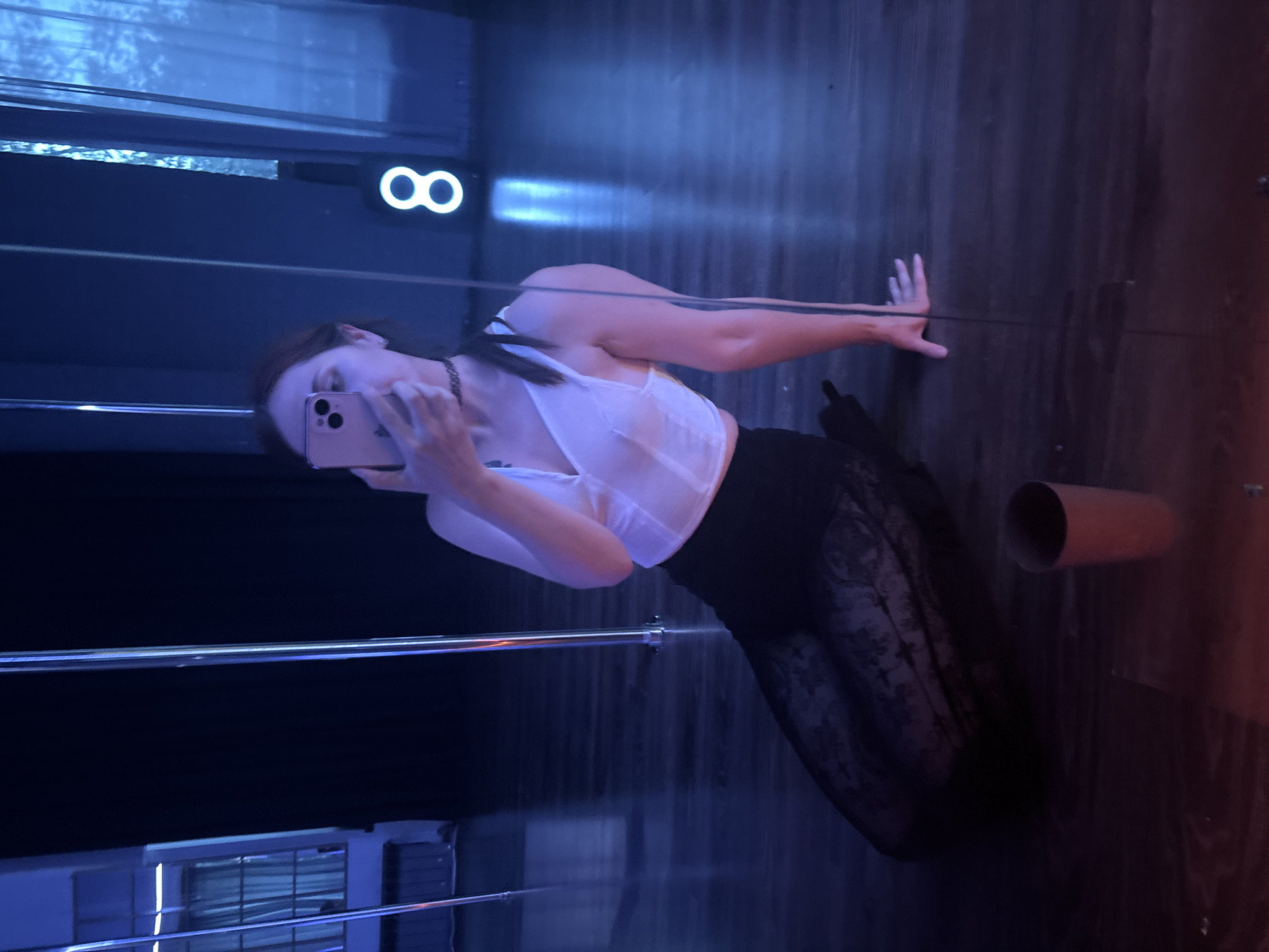 In an indoor studio with polished wooden floors and blue ambient lighting, a person is captured in a selfie. The individual is wearing a white corset-style top and a black lace skirt, and is sitting on the floor. They are holding a light pink smartphone to take the photo. To the left of the person, a metal pole is visible, and to the right, a neon sign displaying the number "8" glows in white. A red cylindrical object, possibly a cup, rests on the floor in the foreground. The scene suggests a dance or performance studio.