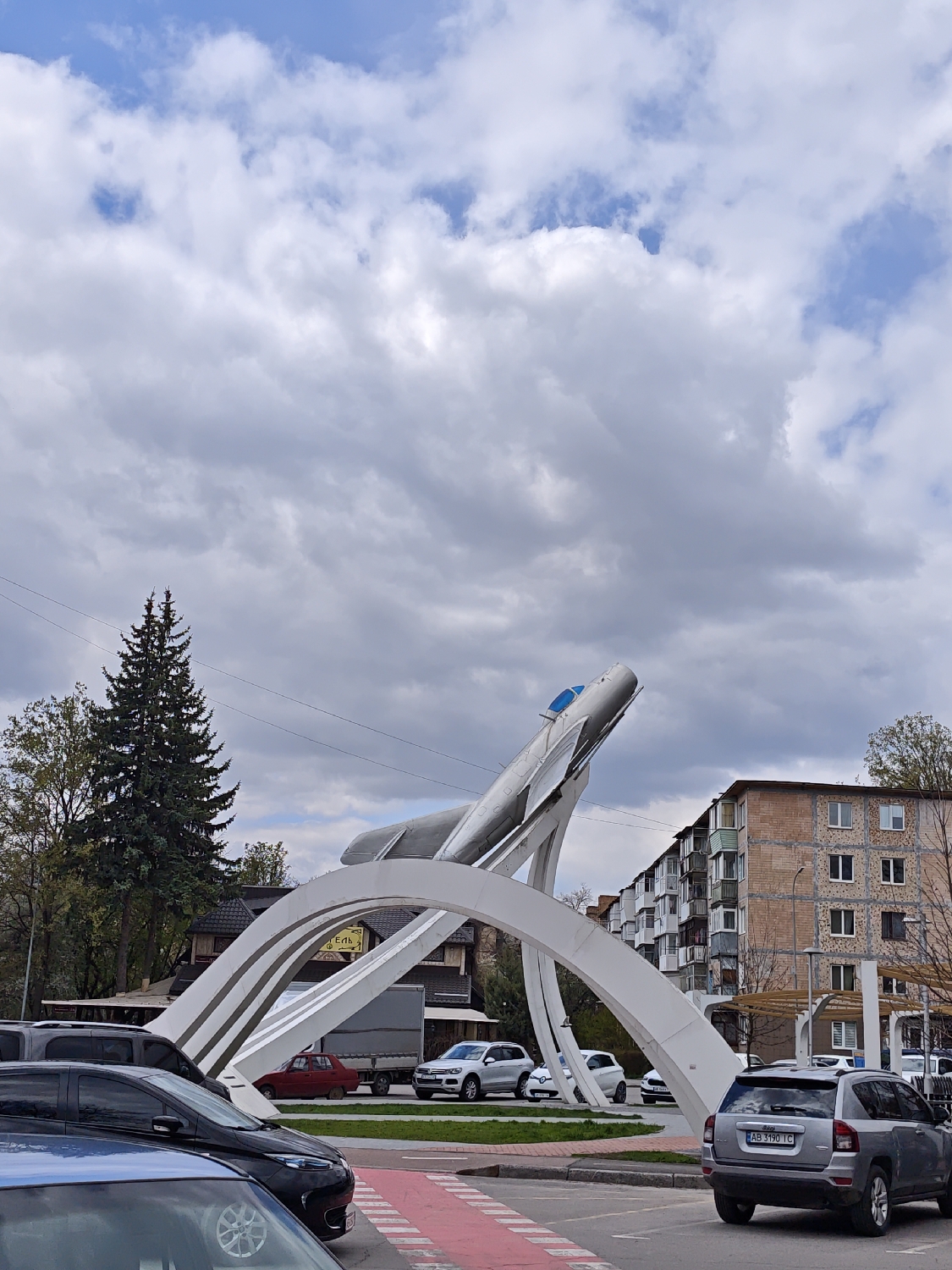 This daytime photograph captures a street scene in Vinnytsia, Ukraine, featuring a prominent aircraft monument under a sky with large white and grey clouds and patches of blue. The central subject is a silver jet aircraft, likely a MiG fighter, mounted dynamically on a series of white, arched support structures, simulating takeoff. This monument is situated in a grassy area, possibly a roundabout or central island.
In the immediate foreground, parts of a black and a blue car are visible. Further back, on the right, a silver/grey Jeep Compass with the Ukrainian license plate "AB 3190 IC" is seen. Other vehicles, including a white SUV and a red car, are also visible in the mid-ground around the monument. A red-painted road marking, resembling a pedestrian crossing or cycle lane, is in the foreground.
To the left of the monument, a tall, dense pine tree stands out, accompanied by other trees. Behind them, a building with a dark roof and a partial view of a yellow sign displaying the text "Erlh" can be seen. To the right of the monument, a multi-story residential building with light brickwork facades, numerous windows, and balconies dominates the background. The scene depicts a blend of urban architecture and greenery, with the aircraft monument serving as a significant landmark.