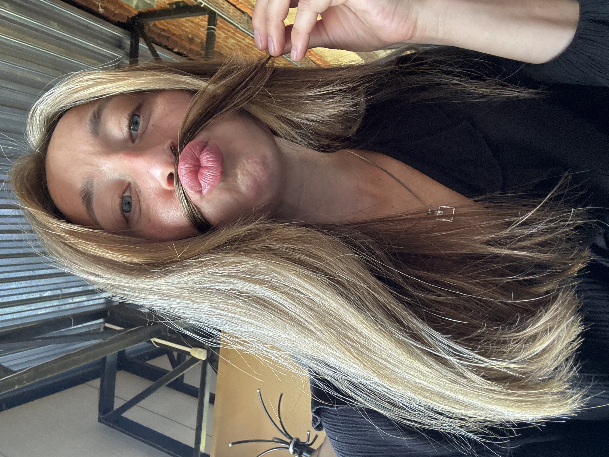 An overhead close-up shot shows a fair-skinned woman with blonde hair posing for a selfie. Her hair is pulled forward, creating a makeshift mustache over her lips, which are puckered. She is wearing a black V-neck shirt with a simple silver pendant. The background is industrial, with exposed metal beams and corrugated metal roofing. The scene appears to be indoors. There is no indication of the city of Chaikovsky, Russia in the image.
