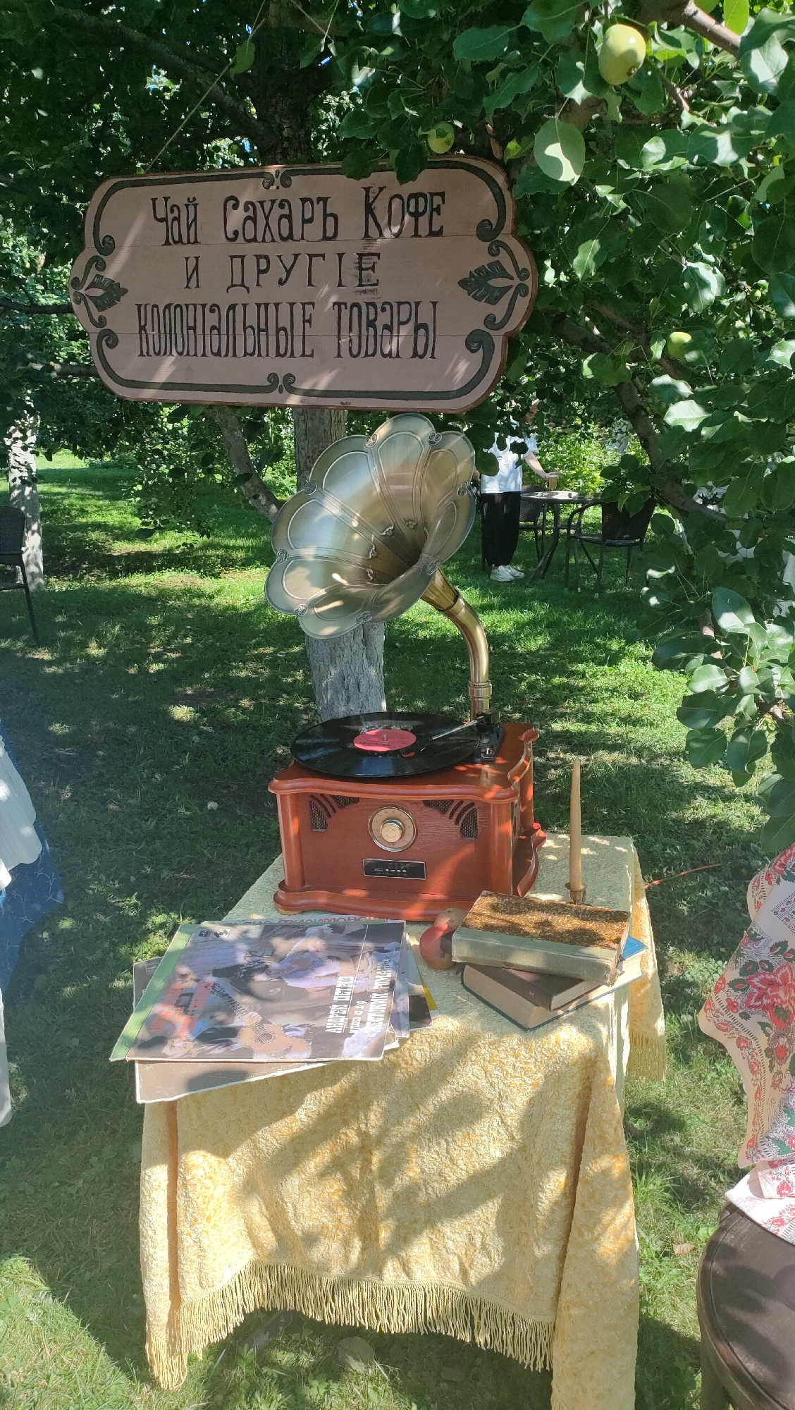 The image depicts an outdoor scene set up as a vintage market stall under a leafy tree. A wooden sign with ornate carvings and Cyrillic lettering hangs from a branch, reading "ЧАЙ САХАРЬ КОФЕ И ДРУГИЕ КОЛОНИАЛЬНЫЕ ТОВАРЫ" (Tea, Sugar, Coffee and Other Colonial Goods). In front of the sign, on a table covered with a yellow fringed tablecloth, sits an antique gramophone with a large brass horn and a record on its turntable. Beside the gramophone are stacks of what appear to be vintage magazines or books. Two people are partially visible in the background, one standing near a table and chairs, and another figure on the right side of the frame wearing a colorful headscarf. The ground is covered in green grass, suggesting a park or garden setting. The scene has a rustic, historical aesthetic.