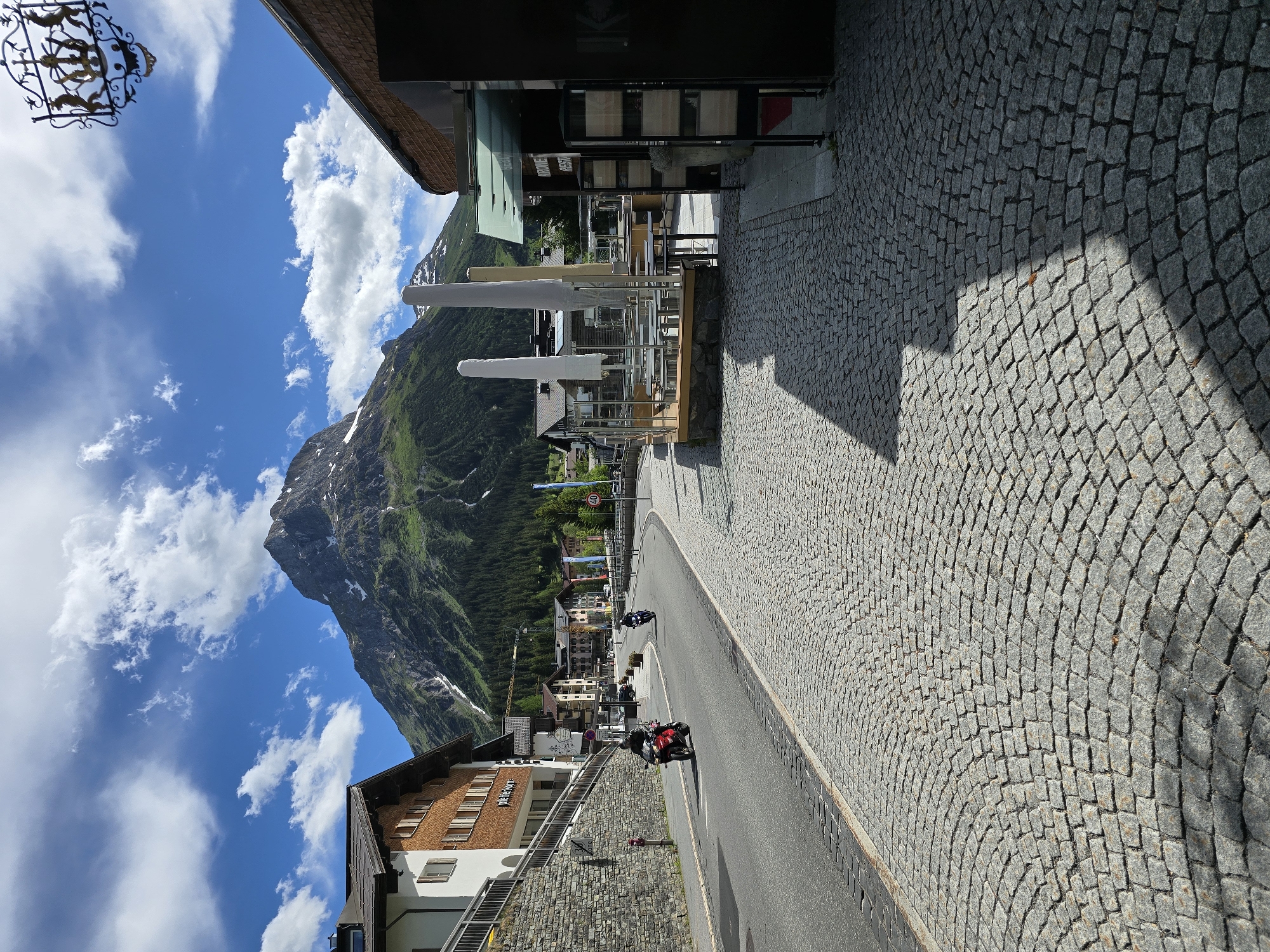 The image depicts a street scene in Lech, Austria, dominated by a large, snow-capped mountain in the background. The foreground features a cobblestone street with a shadow cast by a building on the right. A road curves through the scene, with a red motorcycle in the left lane and a blue motorcycle further ahead. Several buildings line the street, with some displaying outdoor seating areas. The sky is blue with scattered white clouds, and the overall environment suggests a pleasant day in an alpine town.