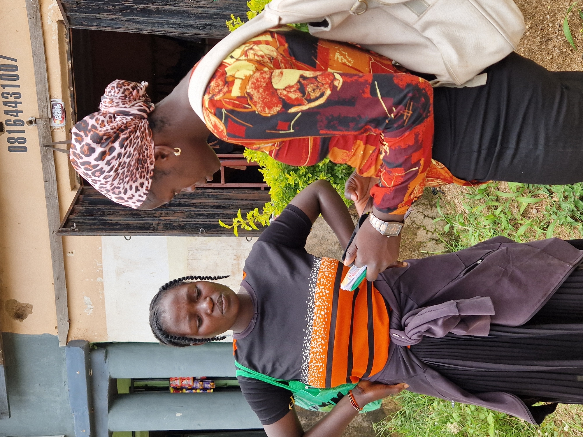 The image shows two women standing outside a building in Kabong, Nigeria. The woman on the left has her hair in braids and is wearing a black t-shirt with orange and white stripes, a dark skirt, and a green bag. She is looking down at a notebook or pad held by the woman on the right. The woman on the right is wearing a patterned headscarf, a colorful long-sleeved shirt, and a black skirt. She has a light-colored backpack slung over her shoulder and is holding the notebook, appearing to be interacting with the woman on the left. The building behind them has a wooden door and windows, with the number "08164431007" visible above the entrance. Green vegetation is visible in the foreground and background.