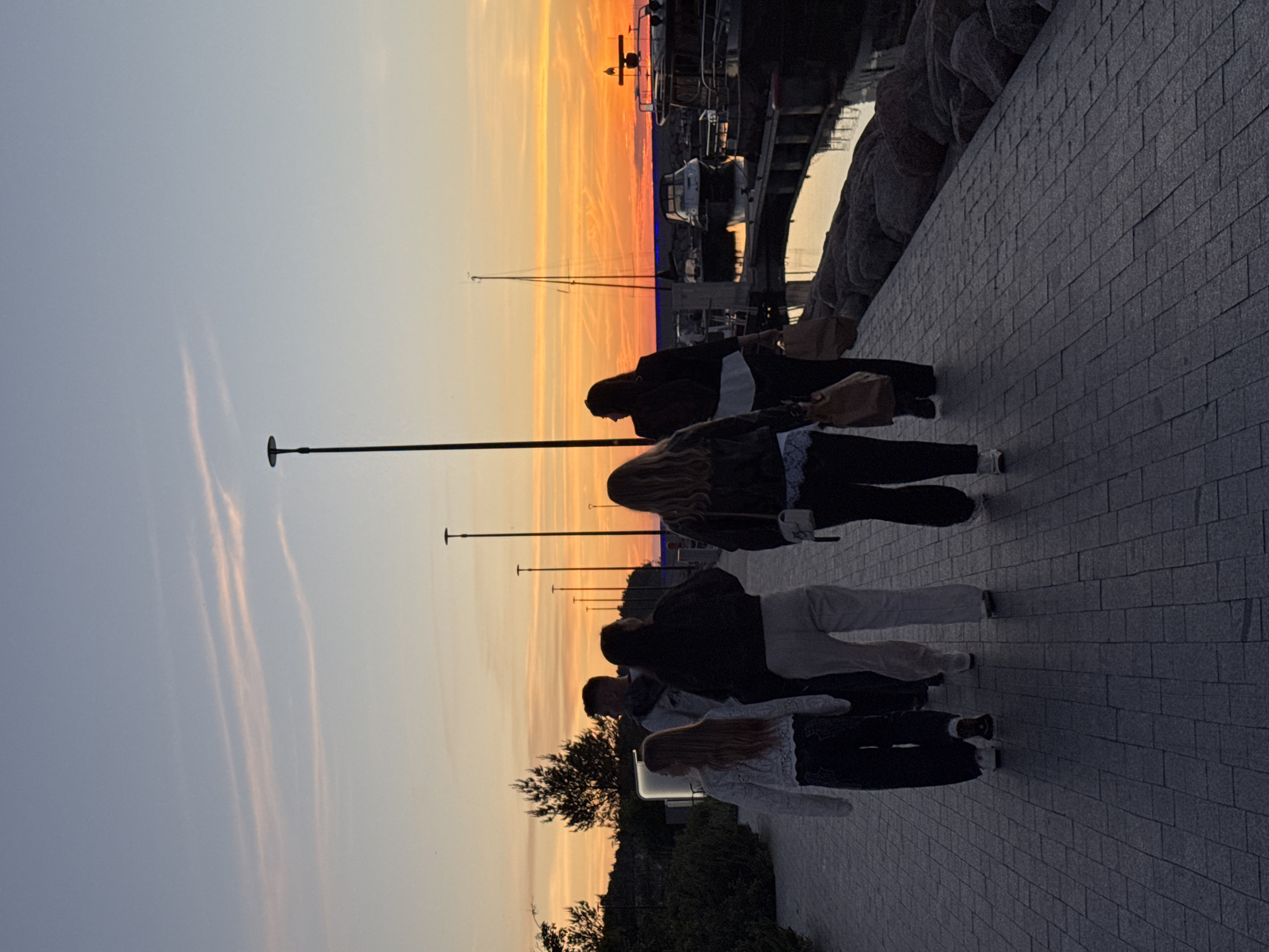 A group of five people, silhouetted against a sunset sky, walk along a paved promenade. The sky is a gradient of orange and pink, with streaks of lighter blue above. There are several lampposts visible along the promenade, and trees line the left side of the scene. On the right, boats are docked in a harbor, with a body of water reflecting the colors of the sunset. The people, whose faces are not visible, appear to be walking away from the camera. Their clothing is casual, and some are carrying bags. The promenade is made of rectangular paving stones, and a low stone wall lines the edge of the water. The overall atmosphere is peaceful and serene. This scene is likely at the Tallinn Harbour area in Tallinn, Estonia.