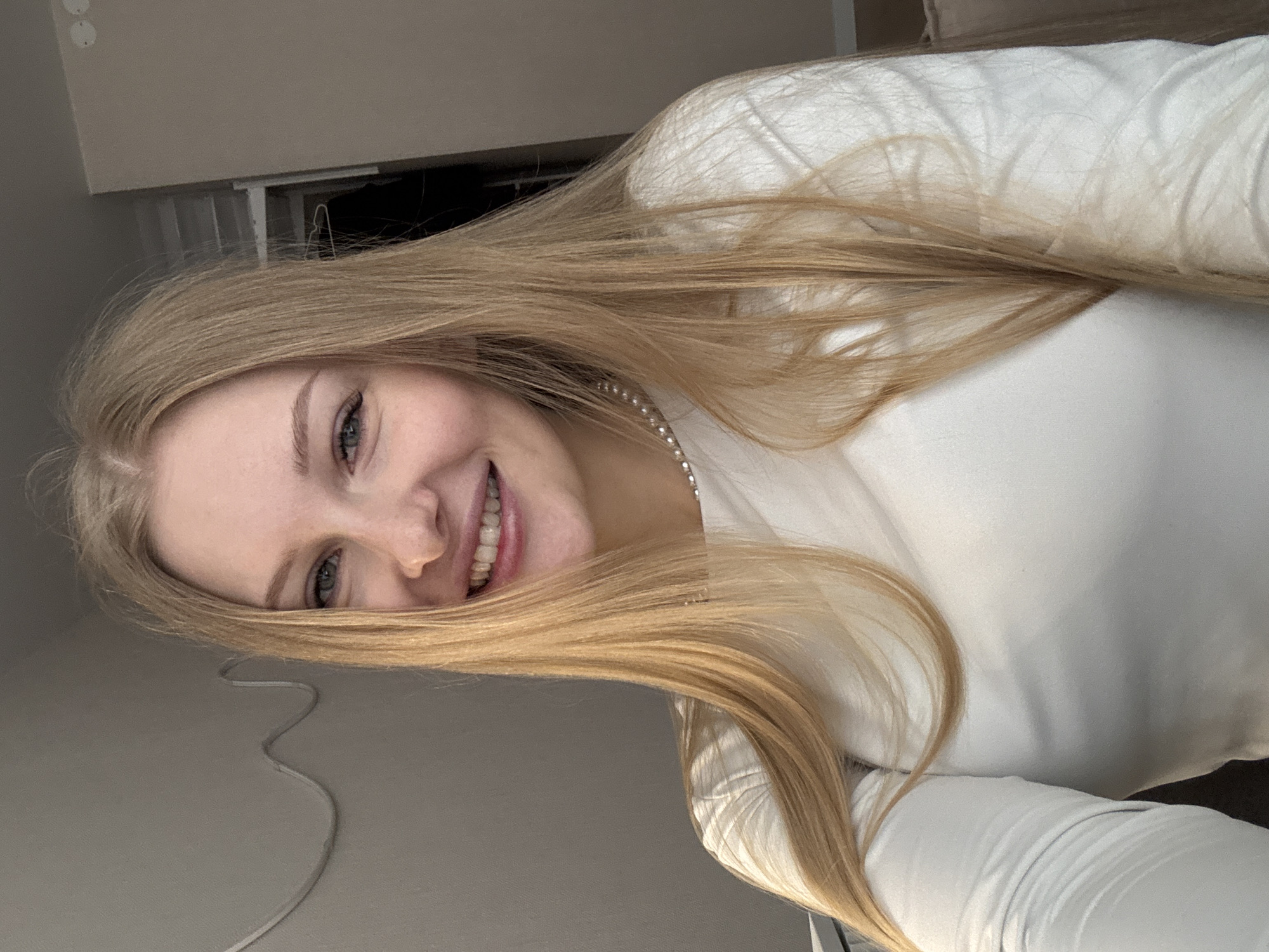 This media file features a young woman in an indoor setting, captured in a selfie-style shot. She possesses long, straight blonde hair, fair skin, and light-colored eyes. Her face is directed towards the camera, and she displays a broad, genuine smile that reveals her teeth, conveying a happy and friendly demeanor.
She is dressed in a plain white long-sleeved top, and a delicate, sparkly necklace, possibly made of small pearls or rhinestones, is visible around her neck. The image is notably rotated counter-clockwise, suggesting a casual pose or an angled camera perspective.
The background indicates an interior space. A light gray wall, featuring two circular wall fixtures (possibly electrical outlets), is visible in the upper left section of the frame. A wavy, light-colored cable rests on this gray surface near her head. Other areas of the background are less defined, showing darker tones that could be furniture or shadowed spaces, along with a contrasting lighter, possibly soft white surface. The lighting is soft and even, characteristic of indoor conditions, providing no specific indicators of the time of day or external weather. No particular activity is depicted beyond the subject posing for the photograph. There is no visible text within the image. The provided location context is Perm, Russia, but this information is not visually verifiable from the image content.