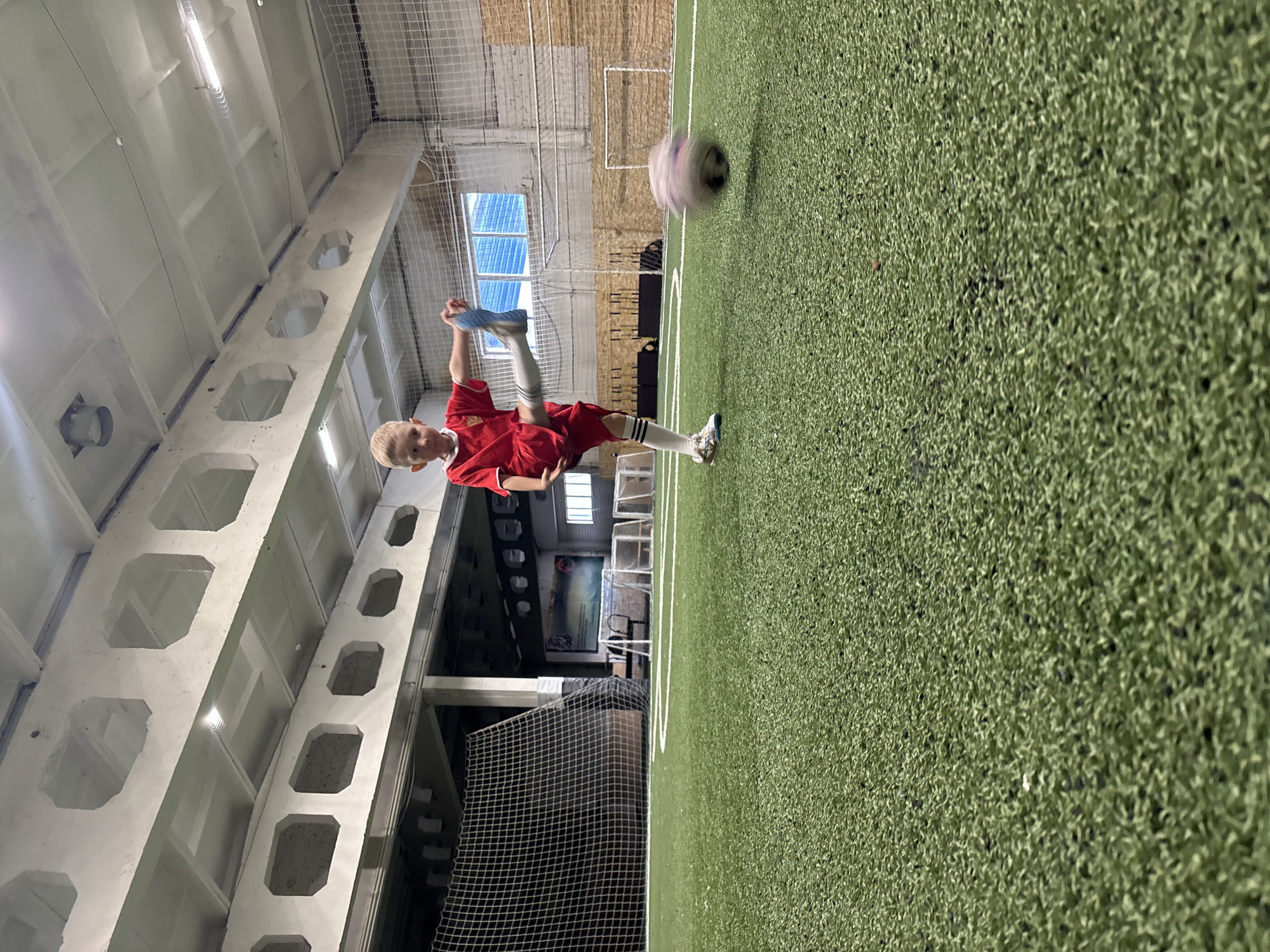 A young boy with blond hair, dressed in a red soccer jersey, black shorts, white knee-high socks with black stripes, and athletic shoes, is captured mid-action on an indoor artificial turf soccer field in what appears to be Chaikovsky, Russia. He is in the process of kicking or juggling a soccer ball, with his right leg raised high and his left leg bent, maintaining balance with slightly raised arms. His gaze is intently fixed on the soccer ball, which appears blurred due to its rapid motion, indicating a dynamic play.
The setting is a large indoor sports facility. The floor is covered with vibrant green artificial turf, marked with white lines delineating the playing area. In the background, a soccer goal with a net is visible. The ceiling is high, featuring distinctive light-colored concrete beams with large, hexagonal or octagonal cutouts, typical of pre-stressed concrete construction. Fluorescent lighting fixtures are mounted on the ceiling, illuminating the space evenly. Mesh netting is present along the walls and possibly as part of the goal structure. A large, high-set window with a blue grid is visible on a back wall. No legible text is visible in the image. The scene captures a moment of active youth sports indoors.