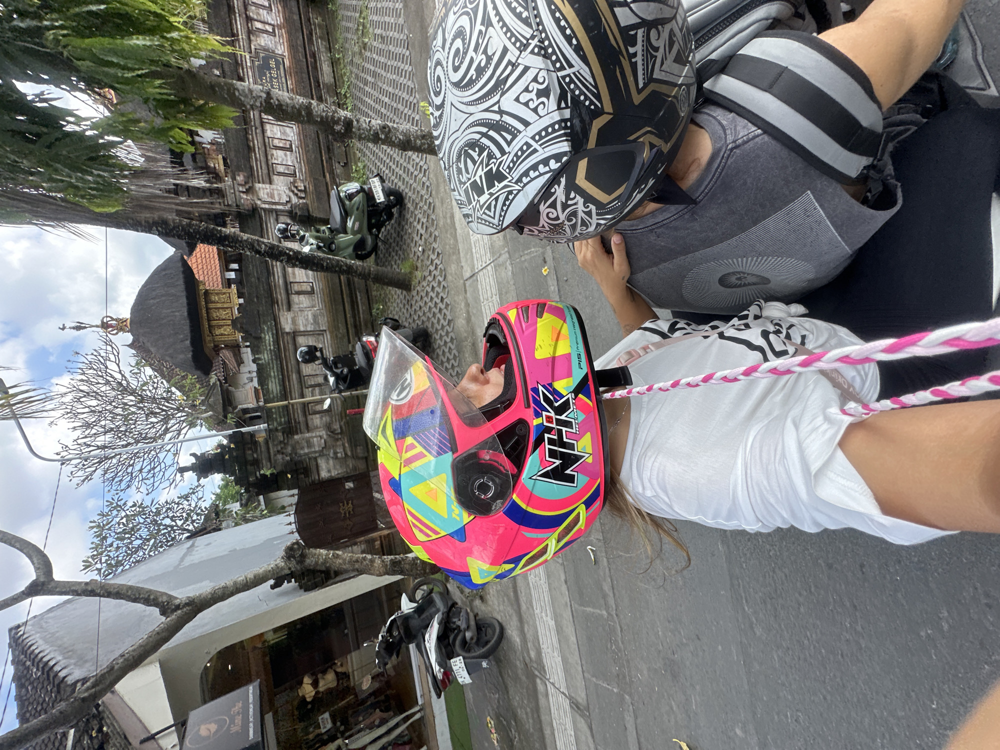Here is a detailed description of the image:
The image shows two people wearing helmets, seated on a motorcycle. The person in the foreground wears a white shirt and a pink and yellow NHK motorcycle helmet. The person behind is wearing a gray shirt and a black and white patterned helmet. The background shows elements of the environment: trees, buildings with traditional Balinese architecture including a roof with thatched layers, and parked motorbikes. A store called "Mime The" is also visible. The image was taken in the city of Mengwi, Indonesia.