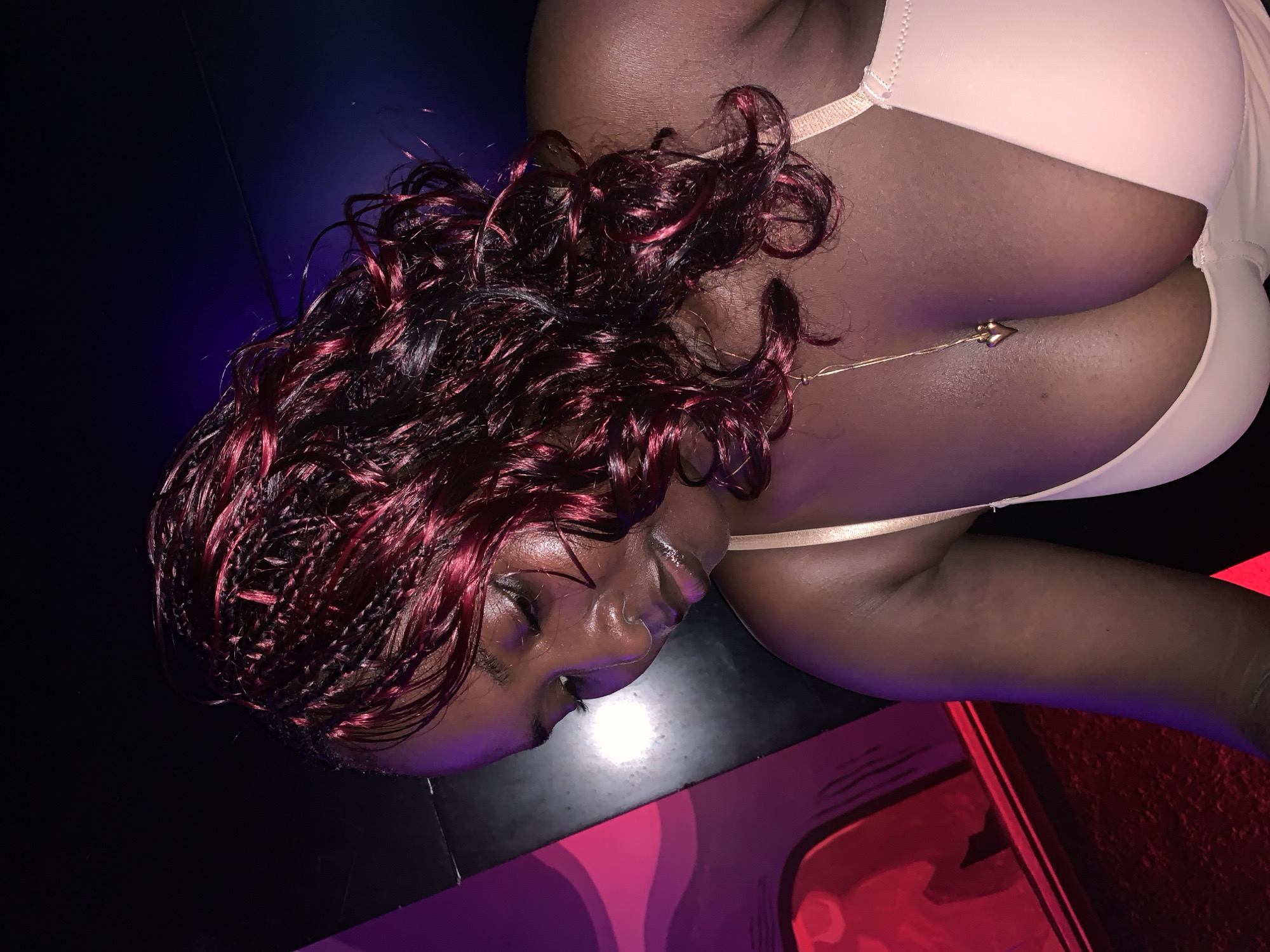A woman with dark skin and burgundy braided hair is seen from the chest up, looking down. She is wearing a light-colored bra and a gold necklace with a heart-shaped pendant. The background is dark with hints of purple and red lighting, suggesting an indoor environment, possibly a nightclub or entertainment venue. There is a purple and pink abstract design on the left side of the frame, which appears to be part of the venue's decor. The image is cropped tightly, focusing on the woman's upper body and hair. There is no indication of location in Howayi, Nigeria.
