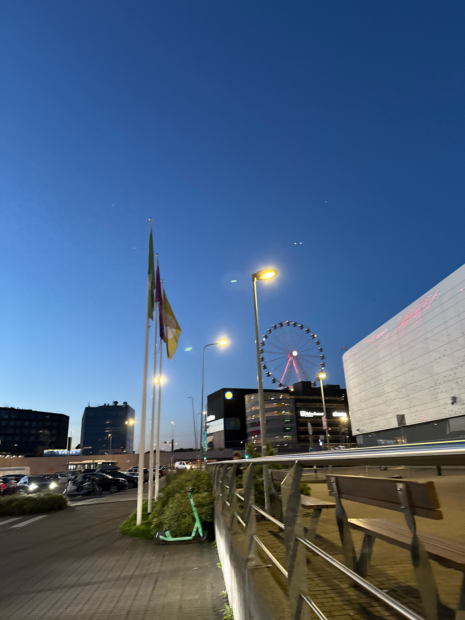 The image depicts a twilight scene in the city of Tallinn, Estonia. In the foreground, a paved walkway is bordered by a metal railing and wooden benches. To the left, a line of parked cars is visible, with streetlights and flags on poles further back. Behind the cars, several modern buildings rise into the clear blue sky. Dominating the mid-ground is a Ferris wheel, its illuminated gondolas a vibrant contrast to the darkening sky. To the right, a large, white, modern building with illuminated red accents stands prominently. A bright yellow streetlight illuminates the area near the Ferris wheel. The overall impression is of an urban environment at dusk, possibly a commercial or entertainment district.