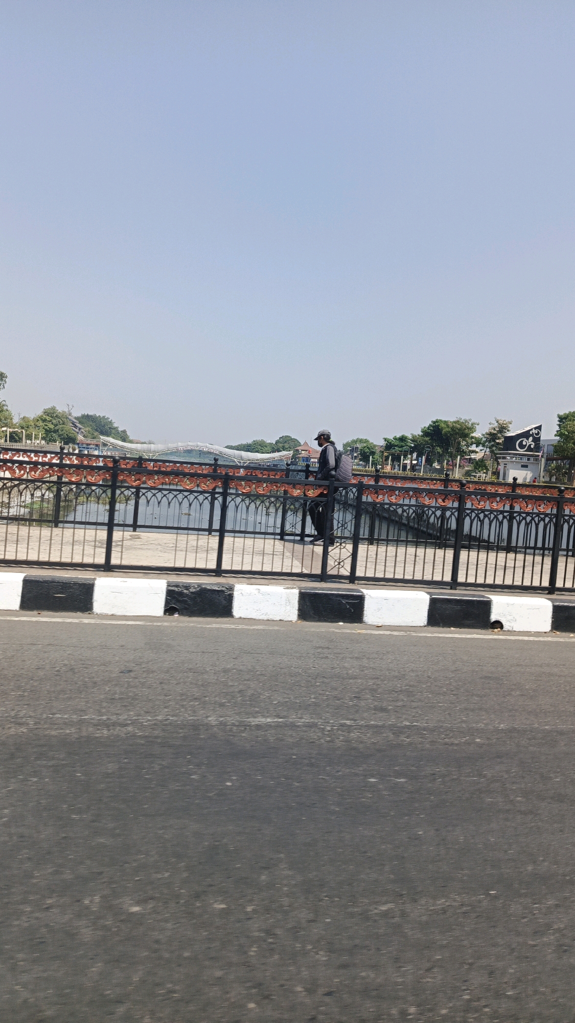 A lone individual wearing a cap, a dark jacket, and dark pants, carrying a backpack, walks along a pedestrian bridge over a body of water. The bridge is characterized by a black metal fence with an ornamental red and orange pattern. In the background, trees and buildings are visible, including a white structure with a black emblem featuring a spiral design. The scene is set against a clear, pale blue sky. In the foreground, a road is marked with alternating black and white bollards. This scene is located in the city of Surakarta, Indonesia.