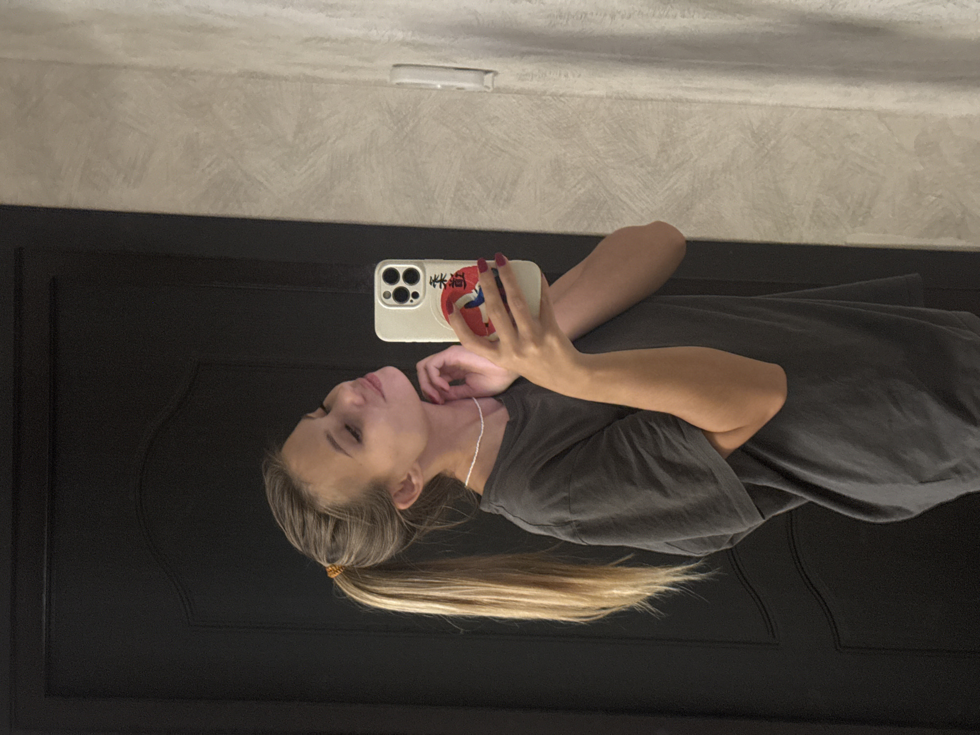 A young woman with blonde hair pulled into a high ponytail stands in front of a dark wooden door, taking a selfie in a mirror. She is wearing a dark grey t-shirt and a thin white beaded necklace. Her right hand holds a white iPhone with a phone case featuring a red and white circular design with Japanese characters. Her left hand is lightly touching her neck. The background to her right is a textured wall. There is no indication of the city of Chaikovsky, Russia, in the image.