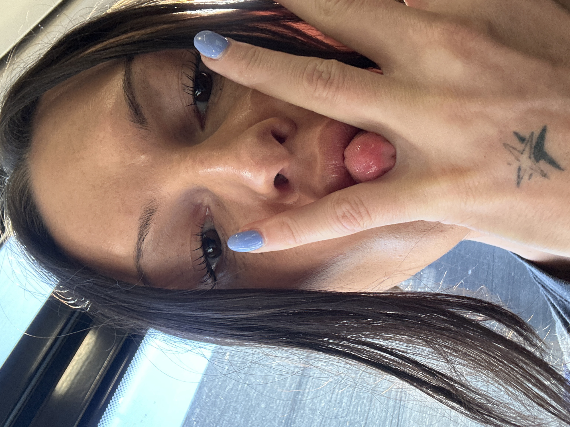 An close-up, angled portrait captures a person, likely female, with dark hair cascading around their face. The subject is seen from a slightly upward angle, with their head tilted. Their left hand partially covers their face, with the index and middle fingers outstretched, forming a "peace" sign or similar gesture, and the thumb and pinky finger obscuring the lower part of their face. Their tongue is sticking out playfully between their fingers.
The person has dark eyes with prominent eyelashes and well-defined eyebrows. Their skin tone appears warm, and there's a visible sheen on their forehead and cheek, possibly from natural oils or lighting. The visible fingernail on the index finger is painted with a light periwinkle or baby blue polish. On the back of the hand, near the wrist area, a small, faded black tattoo resembling an abstract star or burst pattern is visible.
The background is largely out of focus but shows bright daylight coming through what appears to be a window. A dark window frame is visible on the left side, and reflections of the sky or light sources are present. A hint of a dark blue or grey garment can be seen on the right edge of the frame, possibly a shoulder or collar. The overall mood is playful and informal. There is no visible text in the image. Given the provided context, the image was taken in Sevenoaks, United Kingdom, during daytime.