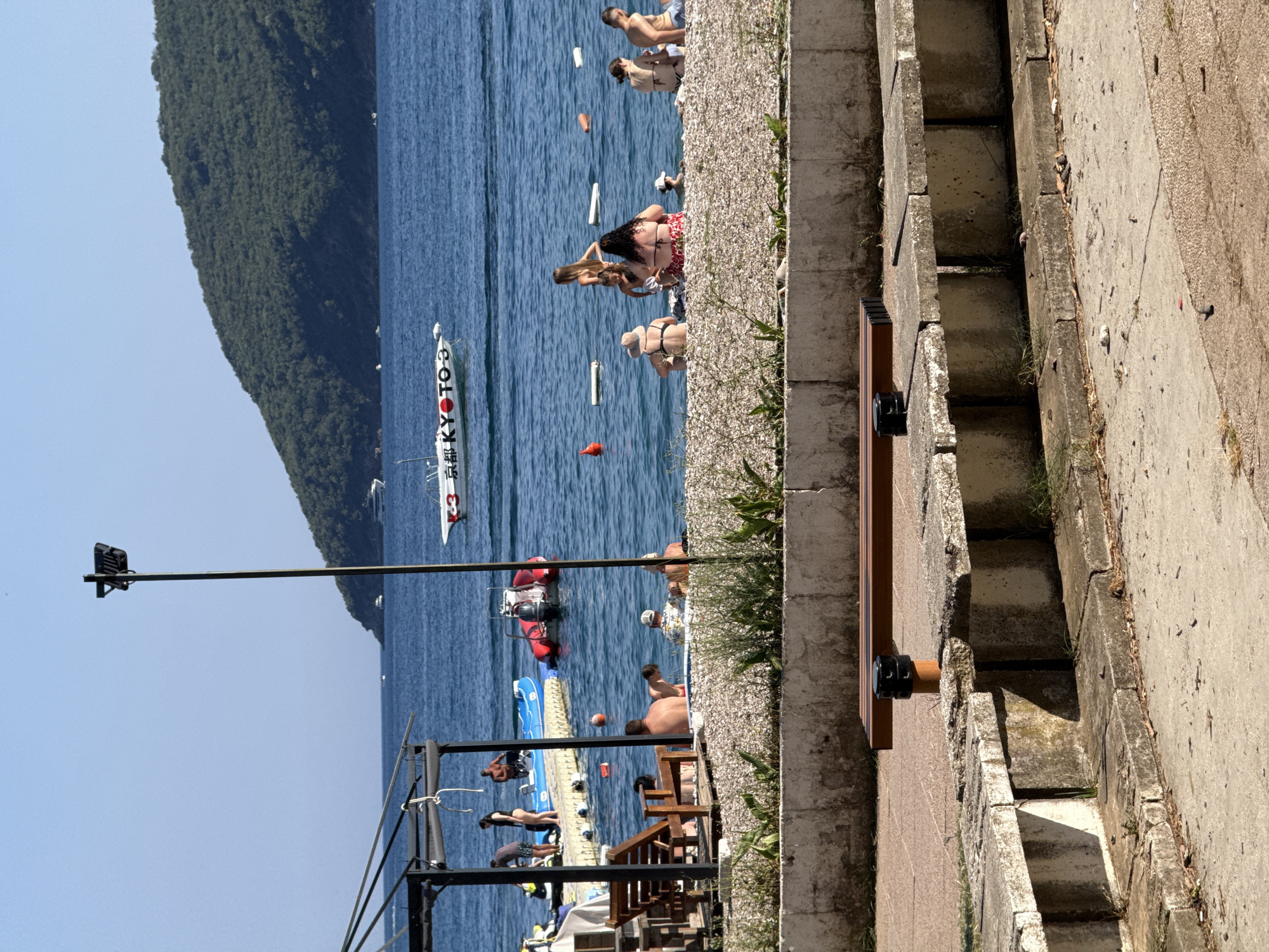 In Budva, Montenegro, a sunny day at the beach is depicted. People are seen relaxing on the pebble beach and in the clear blue sea. Several boats are docked or cruising in the water, including a white speed boat with Japanese writing and the numbers "KYOTO 3" on its side. In the foreground, a concrete bench and a wooden bench are visible, suggesting a promenade area. The background features a lush green, tree-covered hill rising from the sea, under a bright blue sky. The scene captures a typical summer day at a popular coastal resort.