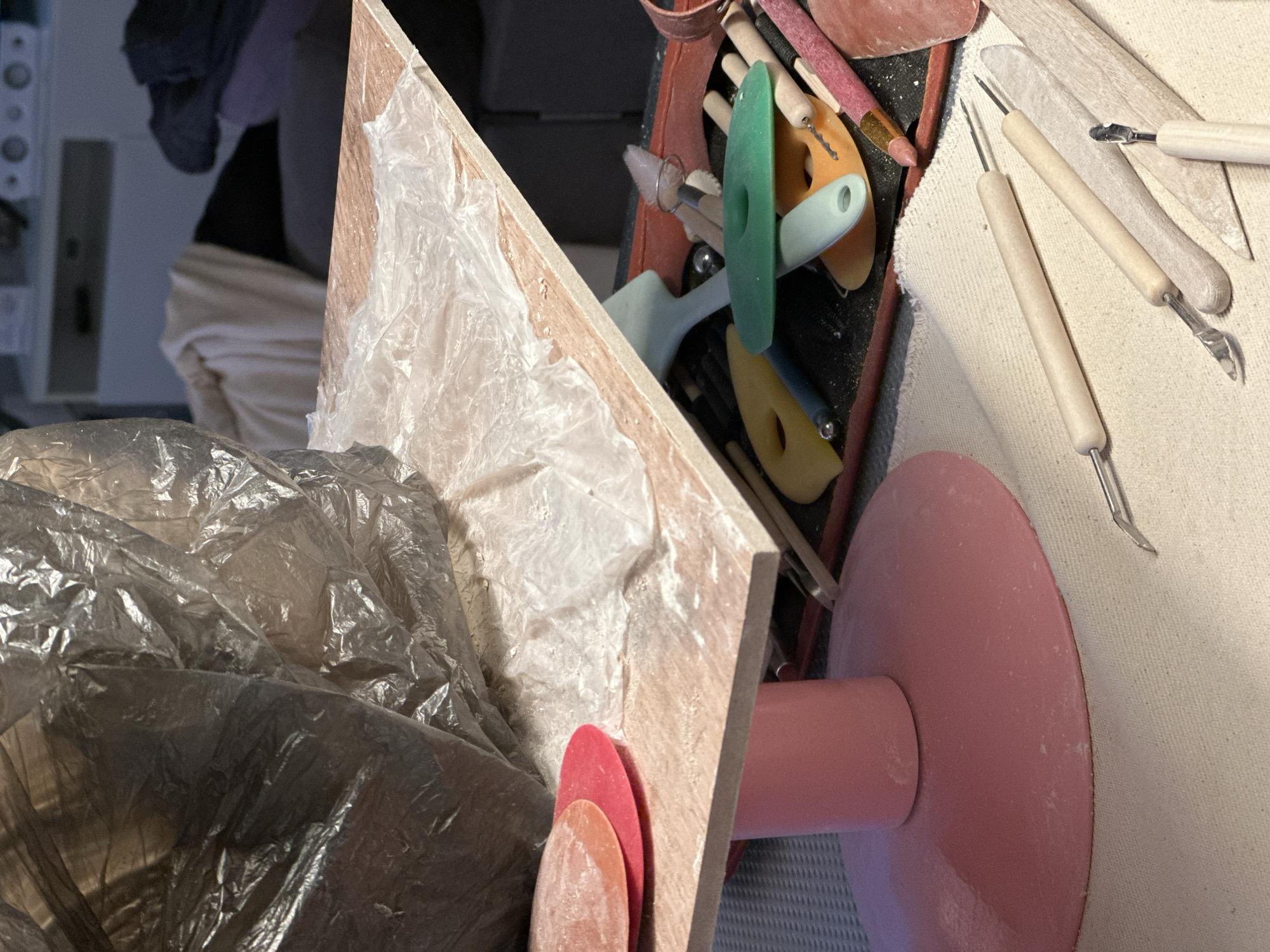 The image depicts a collection of pottery tools and materials laid out on a table, suggesting a crafting or artistic workspace. In the foreground, several wooden-handled sculpting tools with metal tips are visible on a light-colored textured surface, likely a canvas or cloth. To the left, a pink pedestal supports a light-colored, rectangular surface covered with white powder and a translucent plastic sheet. Two circular, reddish-pink objects, possibly clay molds or smoothing tools, are placed on this surface. Behind this setup, a dark plastic bag is loosely draped over some unseen items. Further back, a tray or box holds a variety of colorful pottery tools, including green, orange, and light blue shaping instruments, as well as smaller metal and wooden tools. The background shows glimpses of furniture and what appears to be a shelf with electronic devices. The scene is indoors, with artificial lighting casting shadows and highlighting the textures of the materials. There are no people visible in the image, and no specific identifiable landmarks that would place this scene in Nevinnomyssk, Russia.