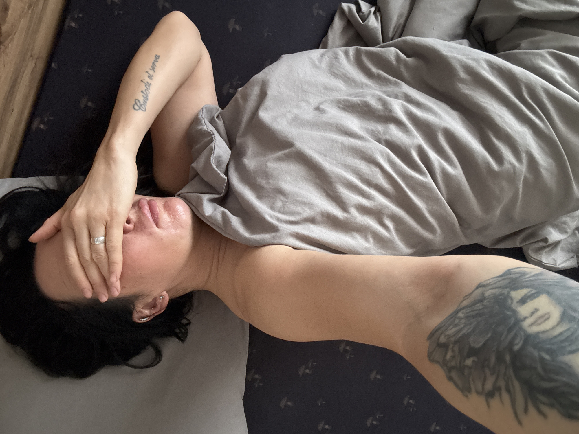 A person is lying in bed, covered partially by a gray blanket. The person has dark hair and is covering their face with their hand, obscuring most of their features. A tattoo is visible on their forearm, and another larger tattoo is visible on their upper arm. The bed has gray and dark blue patterned sheets.
There is no information available to identify the location as Chaikovsky, Russia.