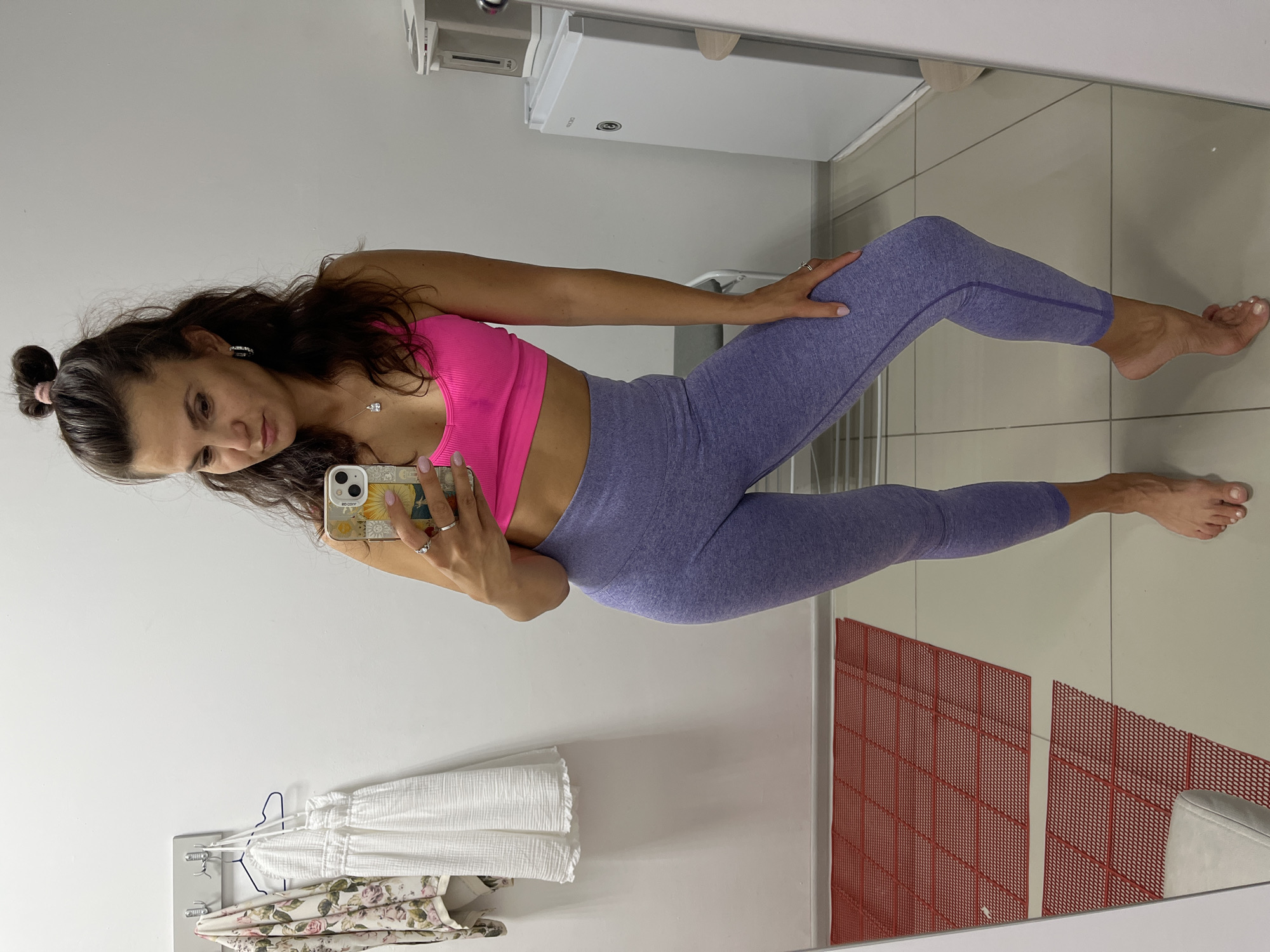 A woman stands in a room, taking a mirror selfie. She is wearing a bright pink sports bra and purple leggings. Her hair is pulled up in a bun. The room appears to be a fitting room or a personal closet, with clothes hanging on a rack and a mirror reflecting the scene. There is a red tiled mat on the floor and a white refrigerator in the background. The image does not provide any information about the city of Petropavlovsk-Kamchatsky, Russia.