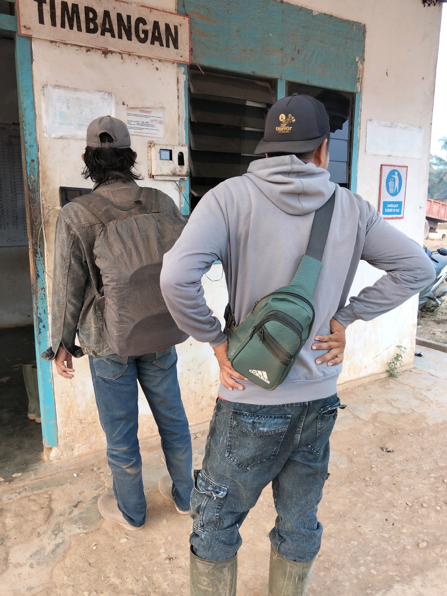 In front of a dilapidated building with a sign reading "TIMBANGAN," two men stand facing away from the camera. The building's exterior is weathered, with peeling paint and exposed wood. The ground is dirt and concrete. The man on the left wears a gray baseball cap, a dark jacket, blue jeans, and light-colored boots. He has a dark backpack. The man on the right wears a black trucker hat with a logo, a gray hoodie, dark distressed jeans, and green rubber boots. He has a green Adidas sling bag across his chest. A trash can symbol with the text "JAGALAH KEBERSIHAN" (Maintain Cleanliness) is visible on the building's wall. The scene appears to be in a rural or industrial area, possibly related to weigh-in services, given the "TIMBANGAN" sign.