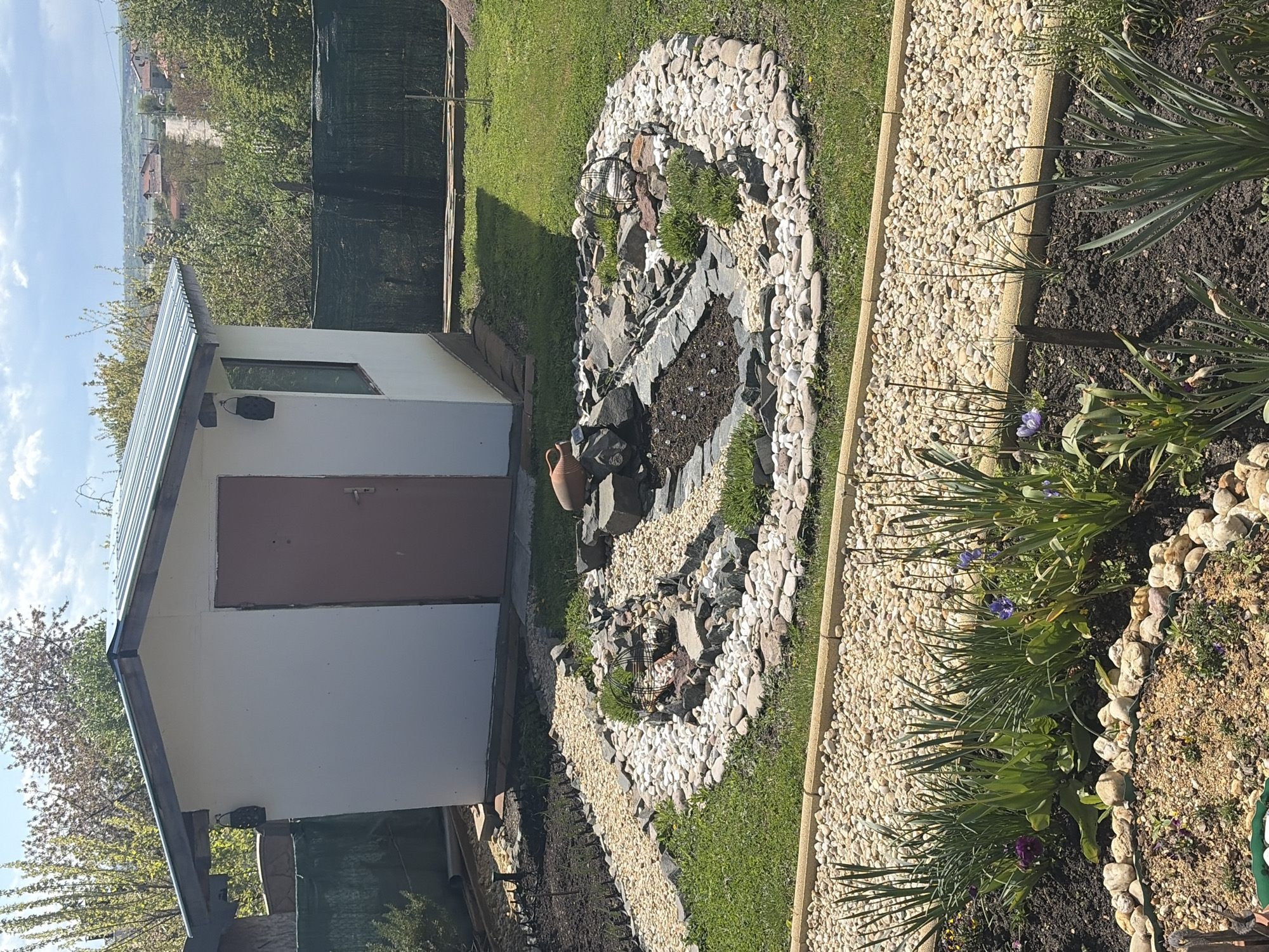 This image shows a garden scene, likely in Konstantinovo, Bulgaria, given the query context. A white shed with a brown door is situated on the left. A decorative rock garden featuring various stones, gravel, and plants lies adjacent to the shed. The garden is interspersed with patches of grass and a gravel path bordered by larger stones. In the foreground, a flower bed contains taller green plants and a few purple flowers. The background includes trees and glimpses of what appears to be a residential area. The sky is partly cloudy.