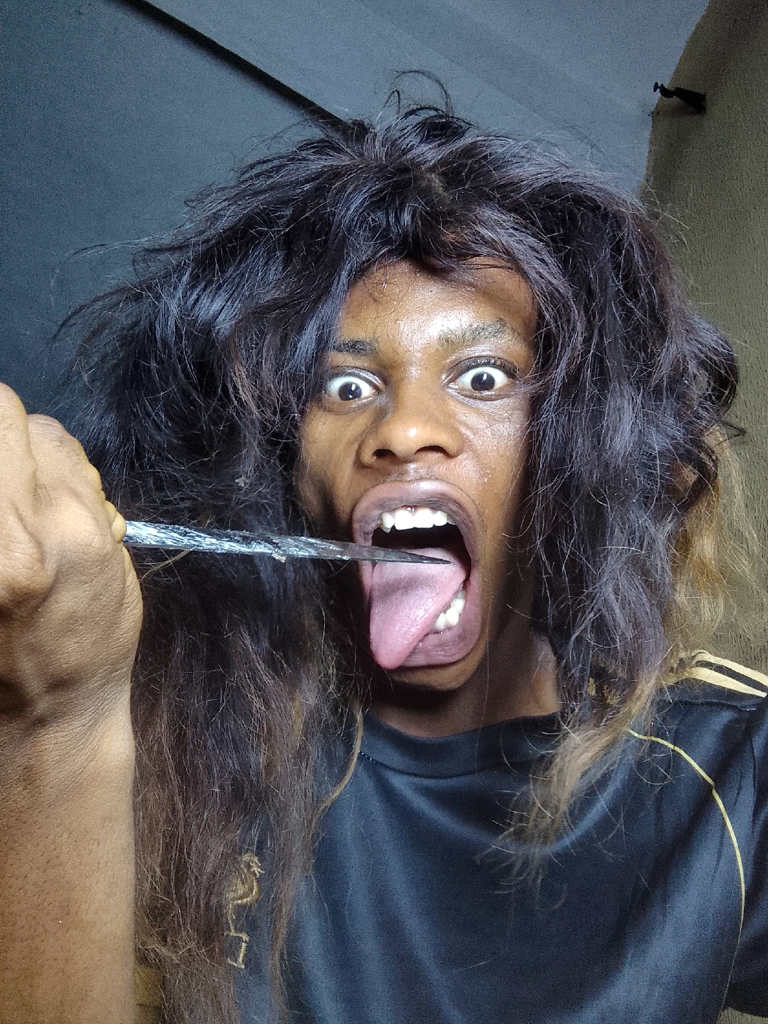A person with dark skin, wide eyes, and dark, disheveled hair is shown holding a silver knife blade parallel to their tongue, with their mouth wide open. The person is wearing a black sports shirt with yellow stripes on the shoulder. The background is blurry and suggests an indoor setting, with gray walls. The city and location are unknown.