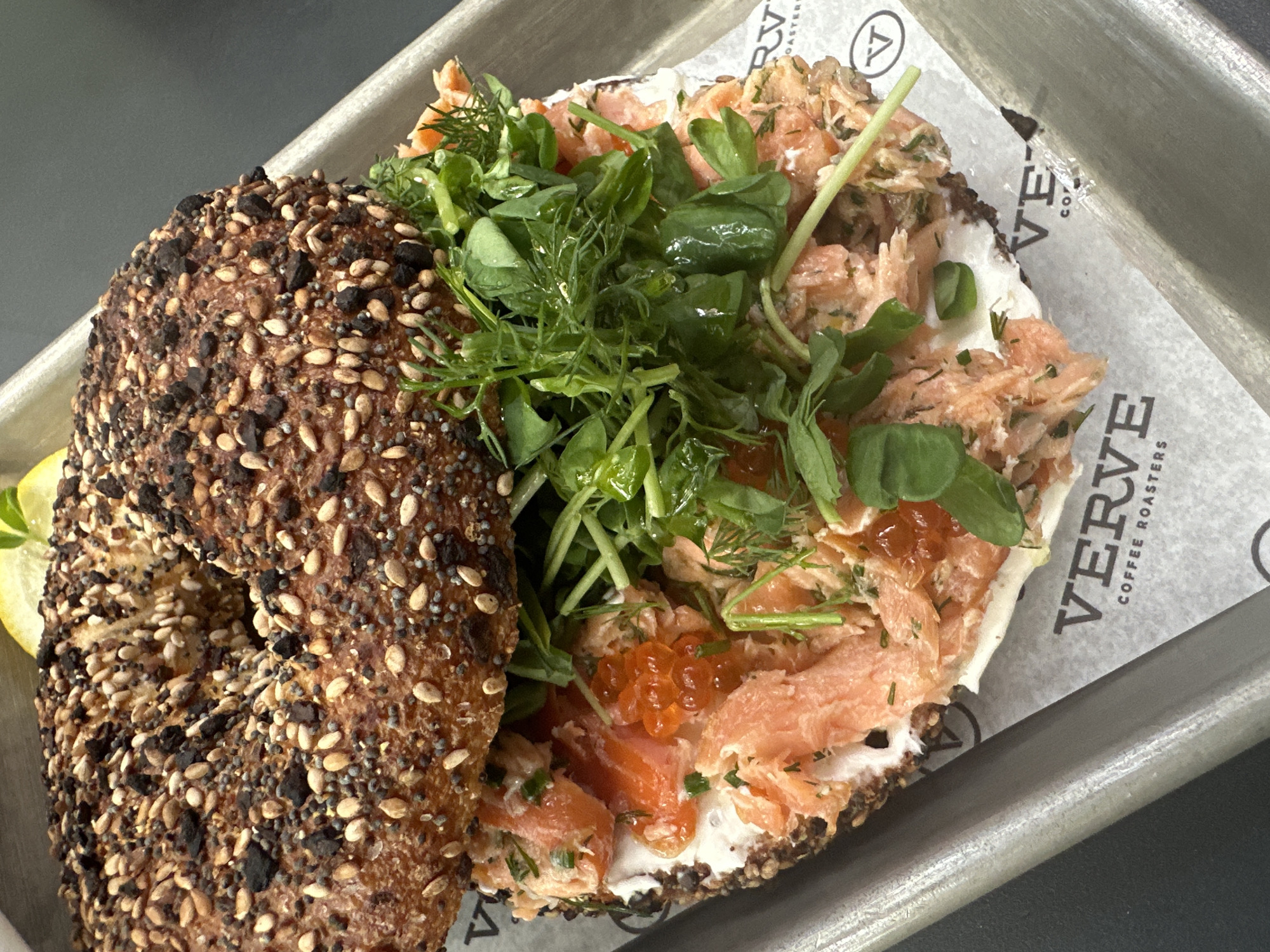 A close-up, high-angle shot captures a gourmet bagel served on a metal tray with a branded paper liner. The bagel, cut in half, is heavily crusted with various seeds, including sesame and poppy, giving it an "everything bagel" appearance. One half is richly topped with a generous portion of smoked salmon or lox, mixed with herbs like dill, and a creamy white spread, likely cream cheese. A cluster of bright orange salmon roe (ikura) adds a pop of color and texture among the salmon. A vibrant pile of fresh green microgreens, possibly pea shoots or a similar delicate herb, is mounded on top of the salmon. A thin slice of lemon is visible on the side, likely intended as a garnish for the bagel. The setting appears to be a cafe or eatery, suggested by the industrial-style metal tray and the branded paper. The paper liner clearly displays the text "VERVE COFFEE ROASTERS" along with a circular logo featuring a stylized "V". The lighting is bright, highlighting the fresh ingredients and diverse textures of the meal.