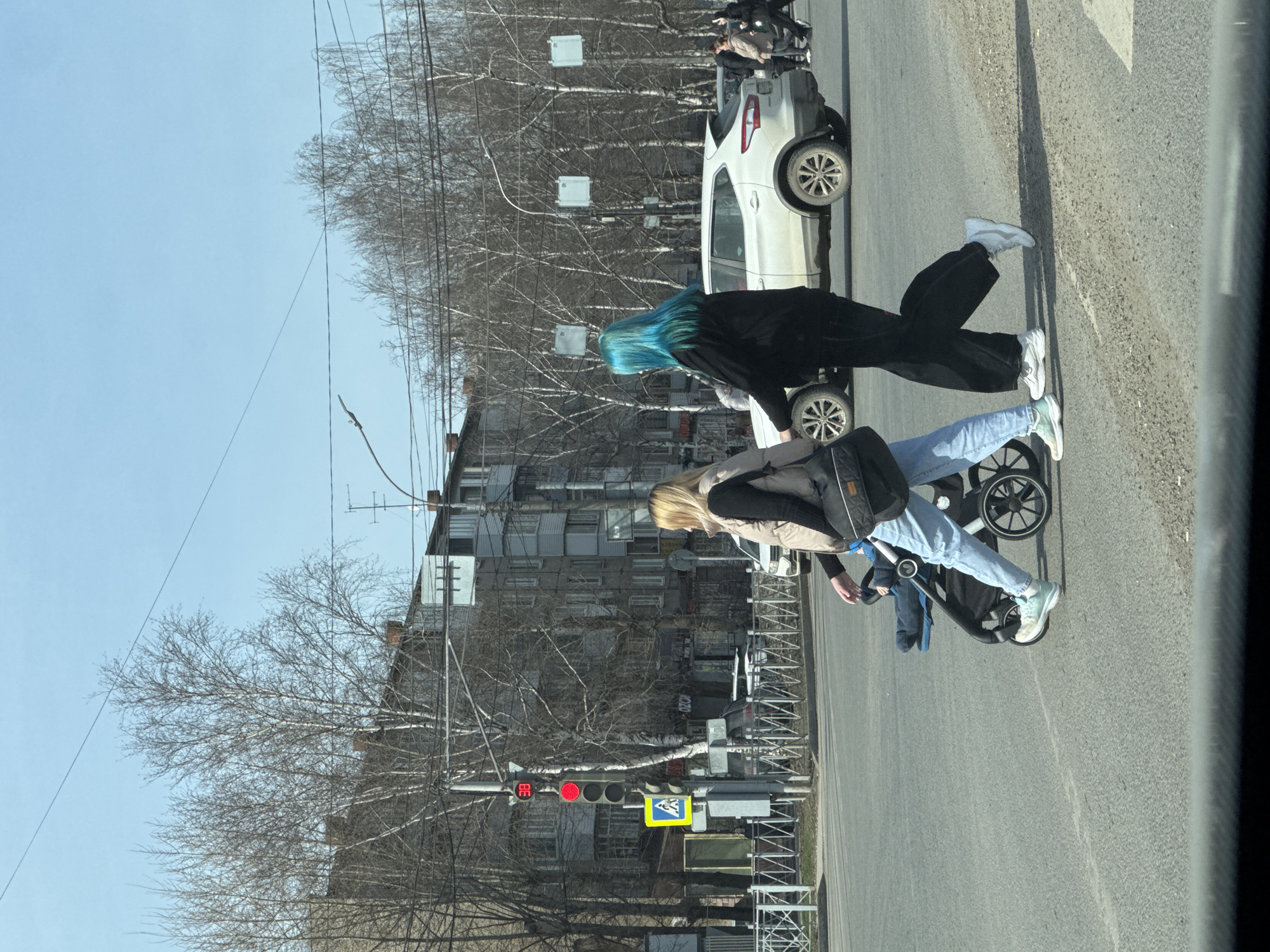 A street scene in Perm, Russia, shows two women and a baby crossing a street. One woman has blond hair and wears a tan jacket over a black shirt, light blue jeans, and light-colored sneakers. She pushes a black stroller containing a baby wearing blue clothing. The second woman has blue hair and is dressed in a black jacket and black pants, wearing white sneakers. They are crossing at a crosswalk with a pedestrian sign. A white car is stopped in the street behind them. A traffic light shows red. Bare trees line the street. In the background is a multi-story apartment building. Street lights and power lines are visible. The photo appears to be taken from inside a vehicle.
