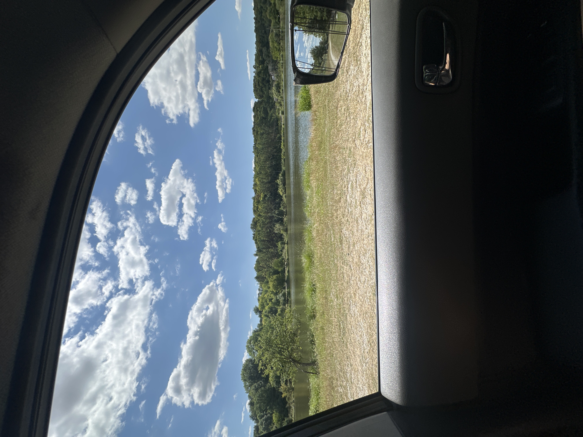 The image is taken from inside a car, looking out the open window towards a body of water bordered by trees under a cloudy blue sky. The foreground shows the car's interior, specifically the dark gray door panel with a chrome door handle. Beyond the car door, a grassy, dry embankment leads down to the water. The body of water is calm and reflects the sky. Across the water, a dense line of green trees forms the natural landscape. A car's side mirror is visible on the right, reflecting a portion of the scene, including trees and sky. The location is identified as Makiv, Ukraine, suggesting this view is from a car parked or driving near a body of water in that city.