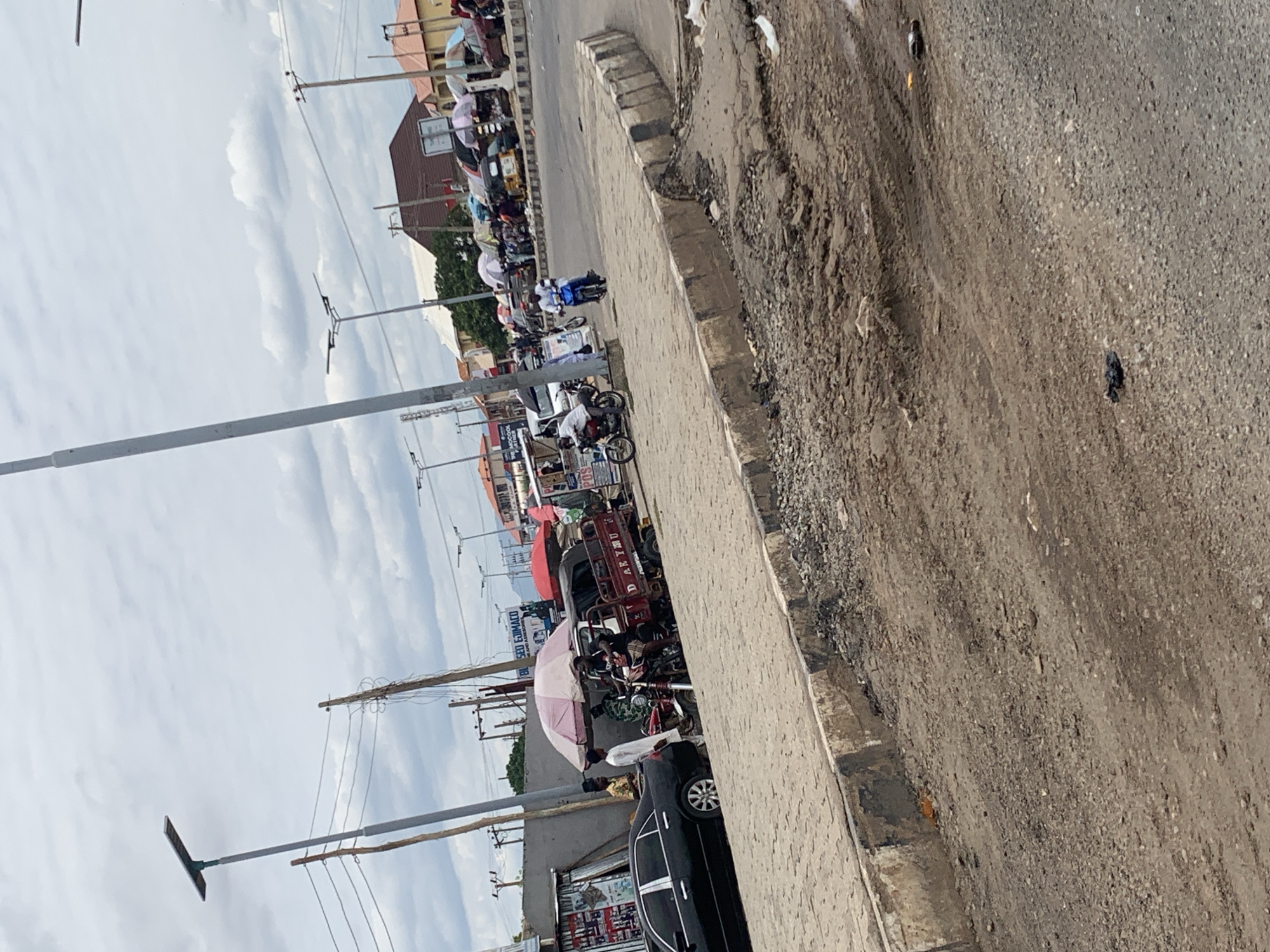 The image depicts a street scene in Lokoja, Nigeria. The view is skewed, angled to the right. A street with pedestrian walkway runs left to right. The walkway borders a road surface with visible damage. Along the left side of the image, various commercial activities are present. People congregate around a three-wheeled vehicle. A black sedan is parked nearby. Above, there are utility poles, wires, and a cloudy sky. In the distance, other buildings and signs are seen.