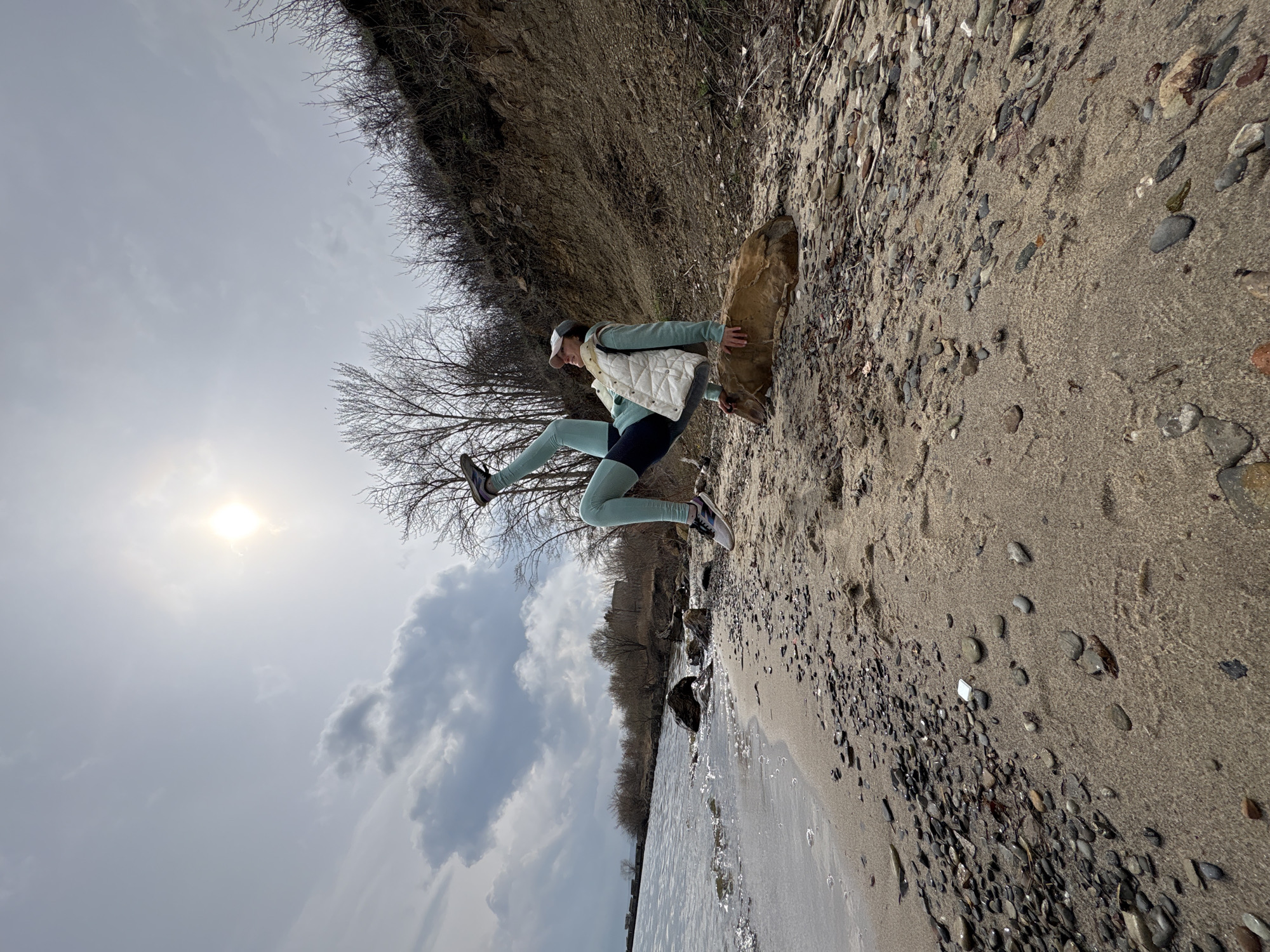 The image depicts a person mid-leap on a beach near water, presumably the Volga River, in Ulyanovsk, Russia, given its location. The subject is wearing athletic attire including leggings, a vest, and a hat, and is positioned near a large rock. The beach is sandy and covered with pebbles, transitioning to a grassy area leading up to a higher ground with bushes and trees. The sky is overcast with the sun partially visible. The water edge is visible in the lower-left part of the frame.