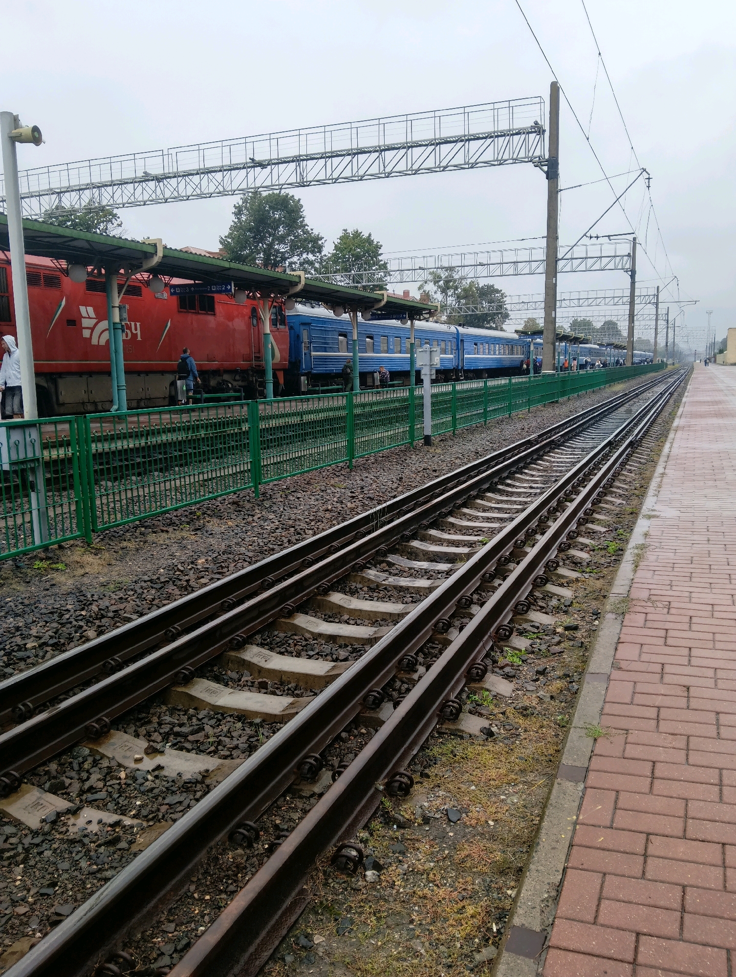 The image shows a train station platform with multiple train tracks and a train. On the left, a red locomotive is coupled to blue passenger cars. The train is on the tracks closest to the platform. To the right of the tracks, there is a paved platform made of red brick. The tracks are ballasted with dark gray gravel and concrete sleepers. Overhead, there are electrical wires and supports for the overhead catenary system. The sky is overcast. In the background, there are trees and buildings. There are a few people on the platform, some near the train and some walking along the platform. The scene appears to be a typical train station environment in Hrodna, Belarus, given the context provided.