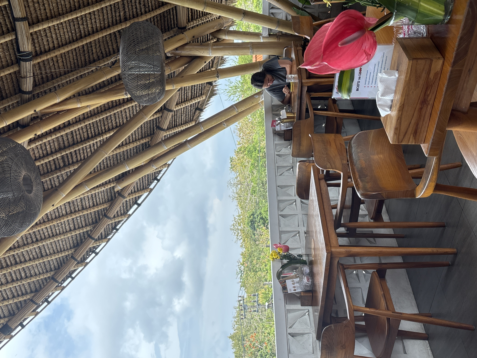 The image shows an outdoor seating area of a restaurant in Kuta Selatan, Indonesia. A person is seated at a table, looking down at their phone. The table is made of wood and has a few items on it, including a glass of water and a tissue box. The seating area is covered by a thatched roof supported by bamboo poles, with hanging lamps. In the background, there are trees and power lines visible, suggesting an urban or semi-urban environment. The overall ambiance appears to be relaxed and casual.