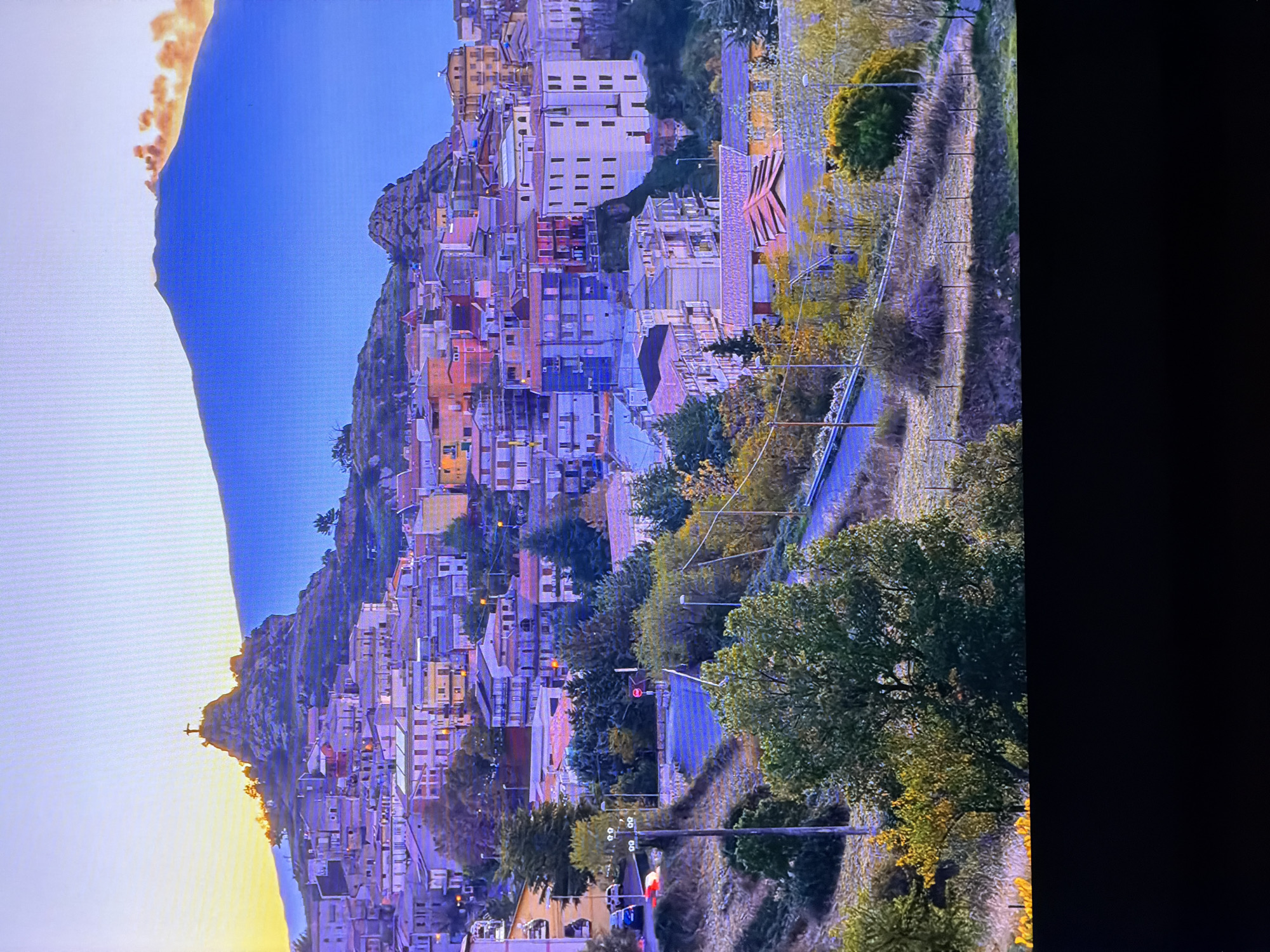 The image depicts a scenic view of a town nestled on a hillside, with a prominent mountain in the background. The town is characterized by its colorful buildings, a mix of residential and multi-story structures, with terracotta-tiled roofs. A notable feature is a rocky outcrop atop a hill within the town, crowned by a statue, possibly of Christ the Redeemer, silhouetted against the sky. The mountain range in the distance is bathed in the warm hues of a sunset or sunrise, with wisps of smoke or clouds tinged with orange suggesting volcanic activity or atmospheric phenomena. The foreground shows a sloped terrain with vegetation, including trees and shrubs, and a winding road. The overall impression is one of a picturesque, possibly Mediterranean, town with a dramatic natural backdrop. The image does not depict the city of Chirchik, Uzbekistan, as its architectural style and landscape are inconsistent with the region.