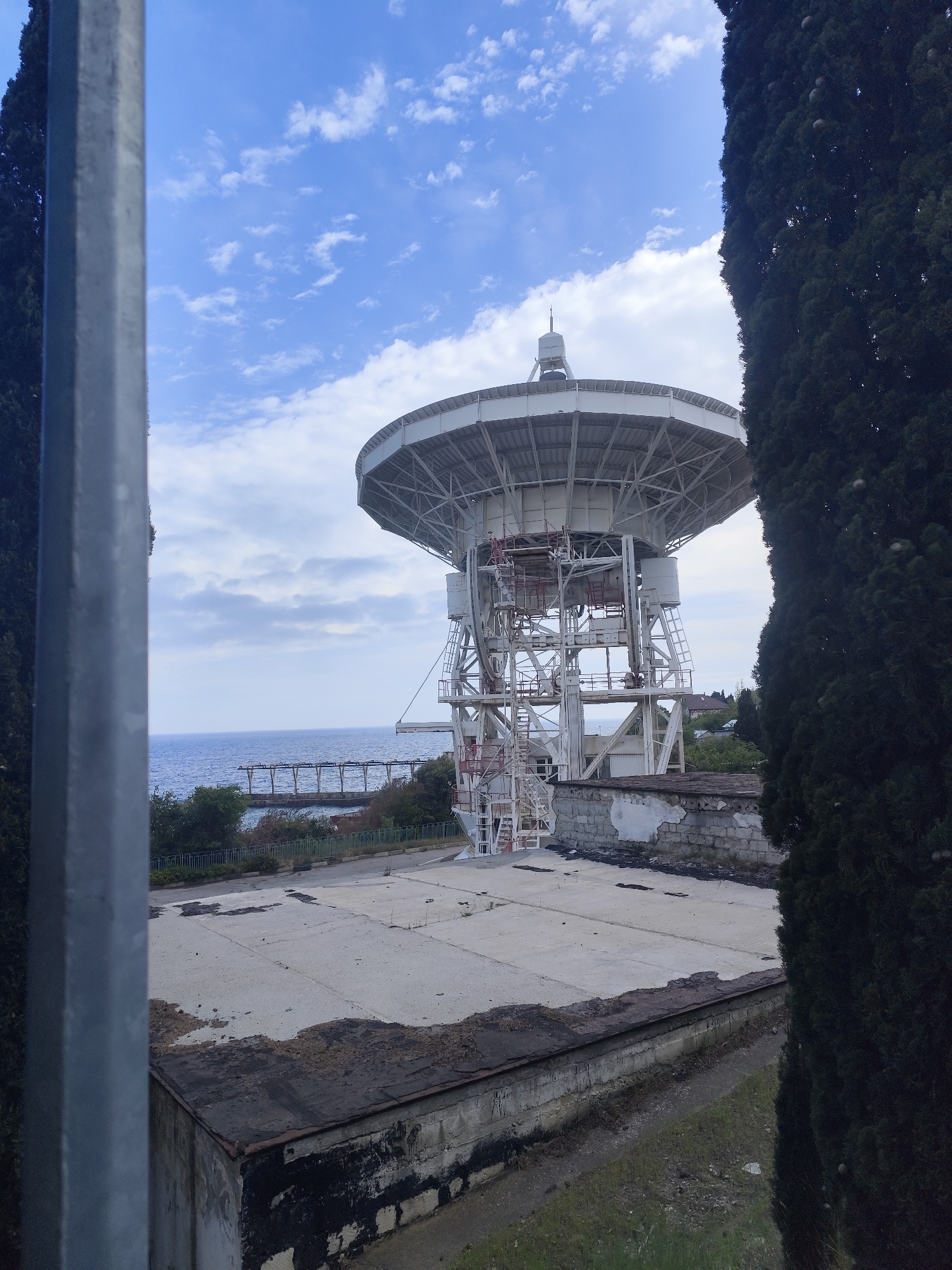 The image captures a large, white radio telescope situated in an area that seems to be either an abandoned industrial site or a research facility in disrepair. The scene is partially framed by a gray metal post on the left and a tall, dark green conifer tree on the right, creating a narrow perspective. The telescope's structure is detailed, with a visible dish and supporting framework, including staircases and platforms.
The immediate foreground reveals a flat concrete surface, possibly the roof of a building, with visible staining and wear. Beyond this area, a glimpse of the sea and a pier can be seen in the background under a sky with scattered clouds. The overall impression is one of abandonment or disuse. The location is identified as being in the city of Unknown, Unknown.