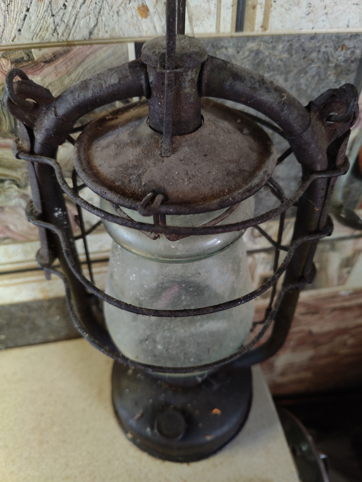 This is a close-up, eye-level, forensic photograph of an old, rusty kerosene lantern. The lantern is dark metal and has a glass reservoir that appears to contain some sediment. It is sitting on a light-colored, textured surface, possibly a table or shelf. The background is out of focus but shows a patterned wall with muted colors and some wooden elements. There are no people present in the image, and no specific actions or interactions are depicted. The overall condition of the lantern suggests it is an old, possibly antique, object. There is no indication of the location in Yarmolyntsi, Ukraine, as the image only provides a visual representation of the object and its immediate surroundings.