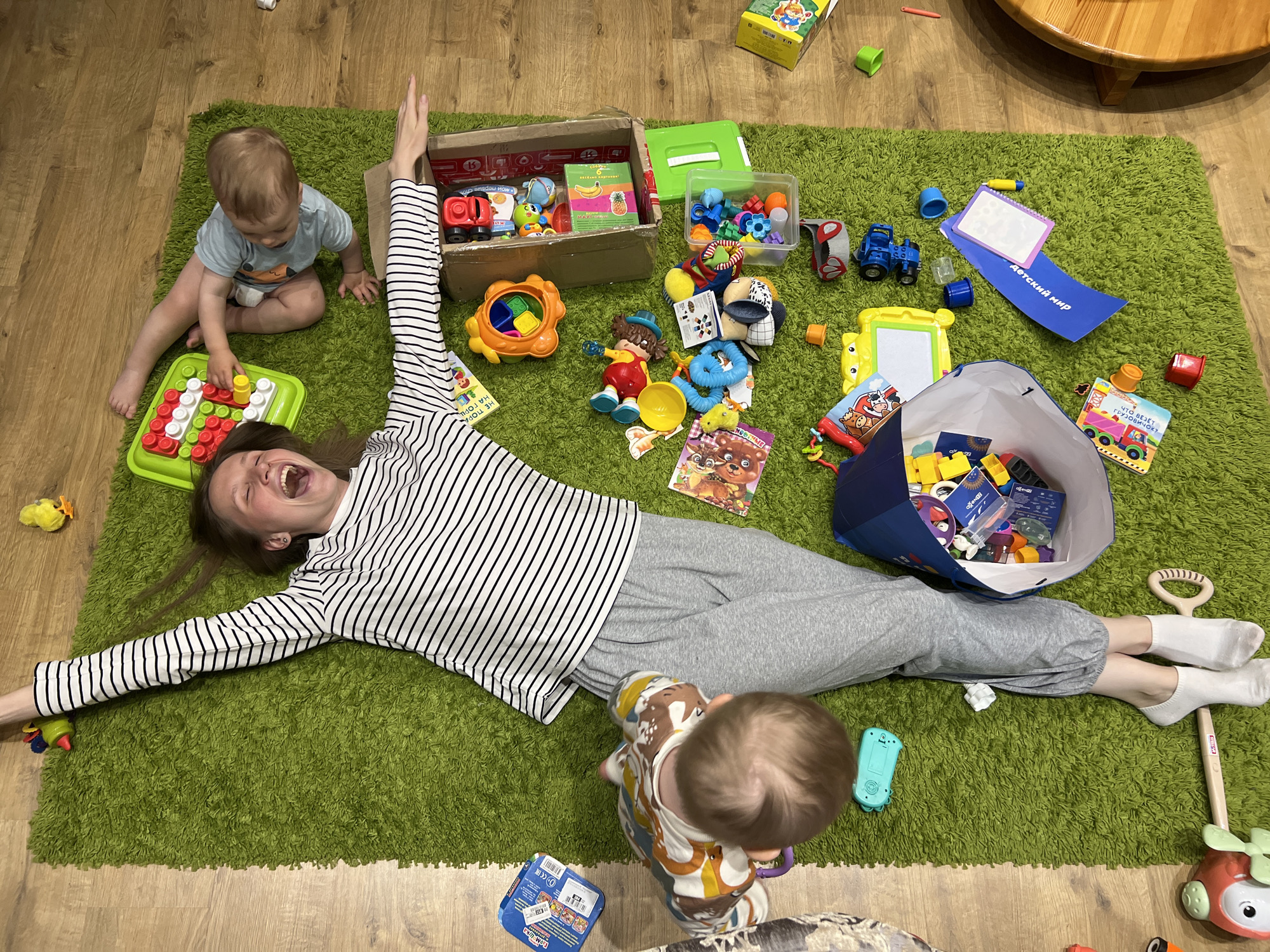 A woman lies stretched out on a green shag rug with her arms extended. She is wearing a striped long-sleeved shirt and grey sweatpants with white socks. She is smiling broadly. Near her legs, a child is standing, and another child sits near her head playing with a toy. Around them are various toys scattered on the rug, including building blocks, toy cars, books, and containers of smaller items. The scene is on a wooden floor. A small part of a table is visible at the top right. The location may be Tomilino, Russia.