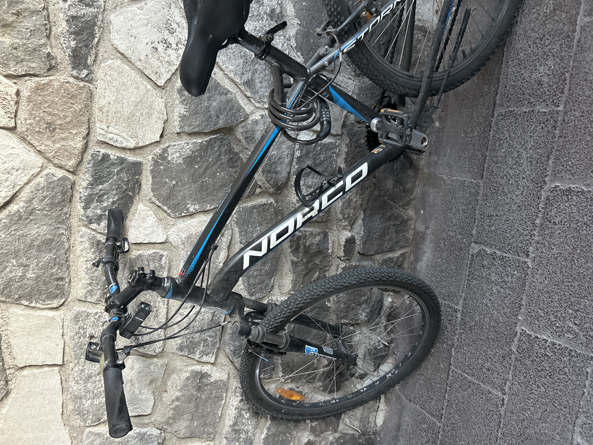 A black and gray Norco Storm bicycle is leaning against a stone wall in an unidentified city. The bicycle is locked with a coiled cable lock. The wall is constructed from irregularly shaped gray stones. The ground near the bicycle shows a darker gray concrete. The bicycle's front tire is near the wall.