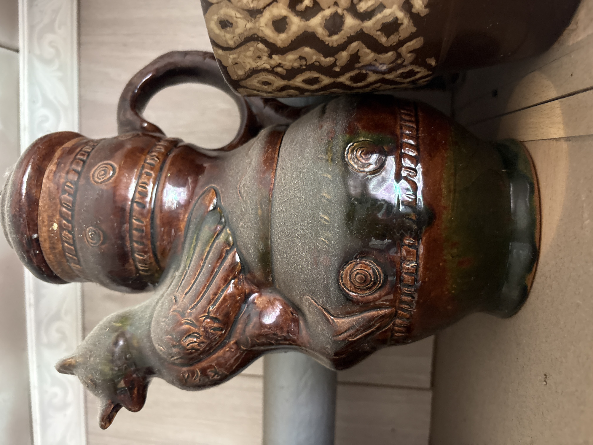 A close-up, eye-level shot shows two ceramic jugs on a shelf in what appears to be a home or storage area in Vetluzhsky, Russia. The jug in the foreground is a dark brown, glazed pitcher shaped like a bird. It has a detailed design with bands of circular patterns and textured lines. The bird's head is to the left, and its body forms the main body of the pitcher. A second jug, partially visible to the right, is also brown and features a repeating geometric pattern. Both jugs appear to be dusty. The background is blurred, with hints of wooden paneling and a metallic pipe.