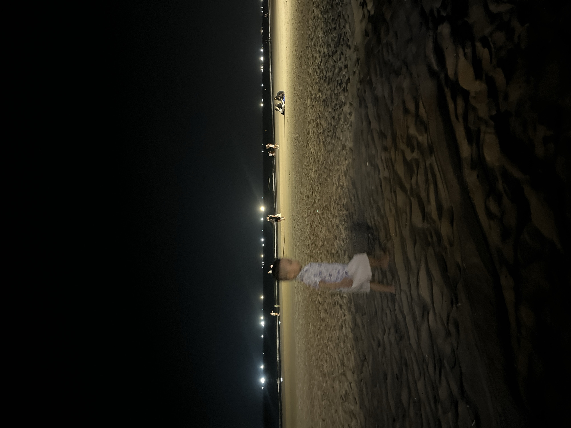 A nighttime scene at a beach in Đà Nẵng, Vietnam. The foreground shows a sandy beach. A child in light-colored clothing stands on the beach, appearing blurred due to motion. In the distance, a line of faint lights indicates the presence of the city or a pathway along the beach. Several individuals are visible in the distance, positioned closer to the lights. The sky above is dark and featureless.