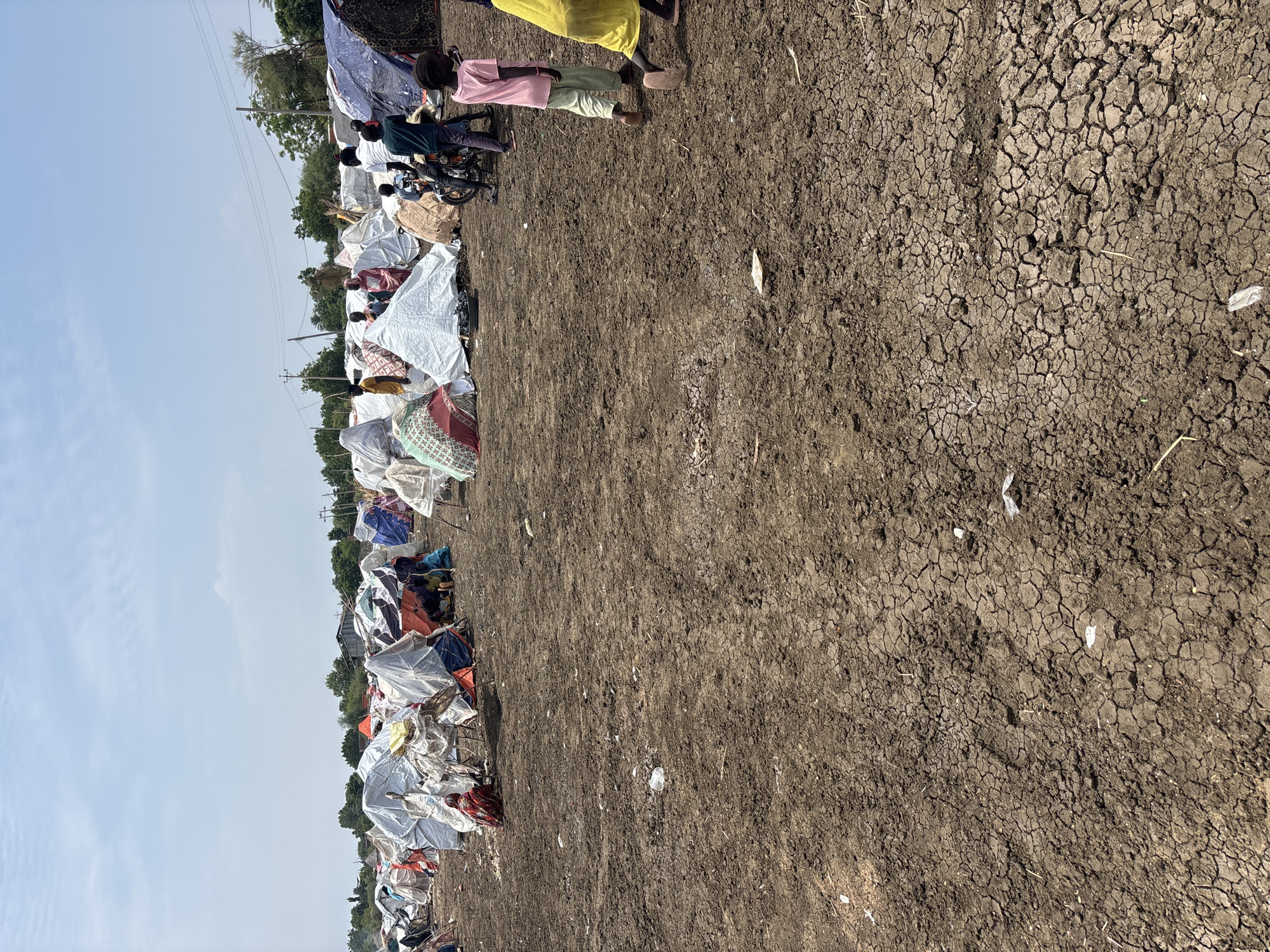 The image shows a refugee camp in Al Babali, Sudan, with numerous makeshift tents and shelters. The ground is dry and cracked, indicating arid conditions. Several people, including adults and children, are visible among the tents. Some are seen walking, while others are gathered near the shelters. The tents are made from various materials, including tarps, plastic sheeting, and fabric, and are set up close together. The overall scene depicts a temporary settlement established in a rural or undeveloped area, typical of displacement situations.